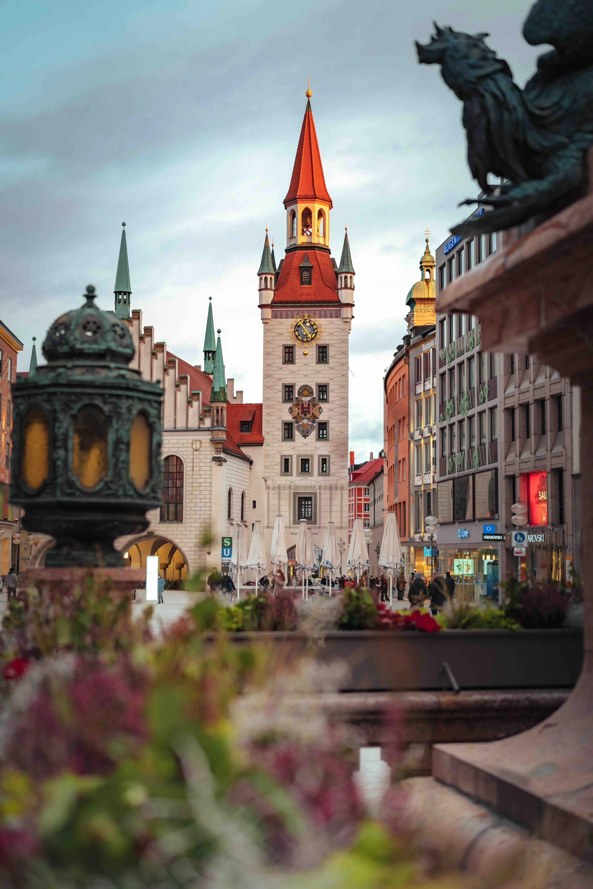 Munich, Germany | Old Town Hall (Altes Rathaus) clock tower on Marienplatz at dusk, viewed through flowering plants in the foreground.