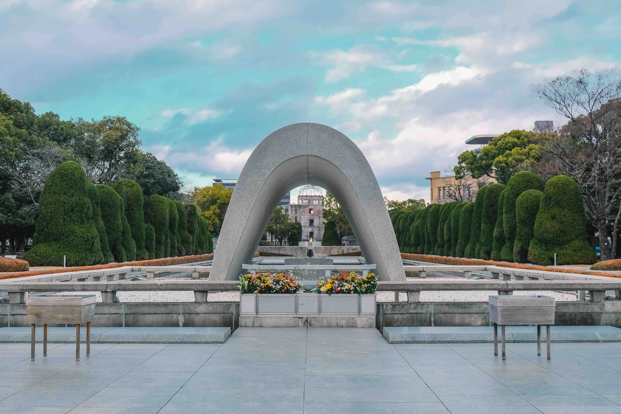 The Hiroshima Peace Memorial cenotaph arch in Peace Memorial Park, framing the Atomic Bomb Dome and the Flame of Peace beyond, with flower offerings in the foreground