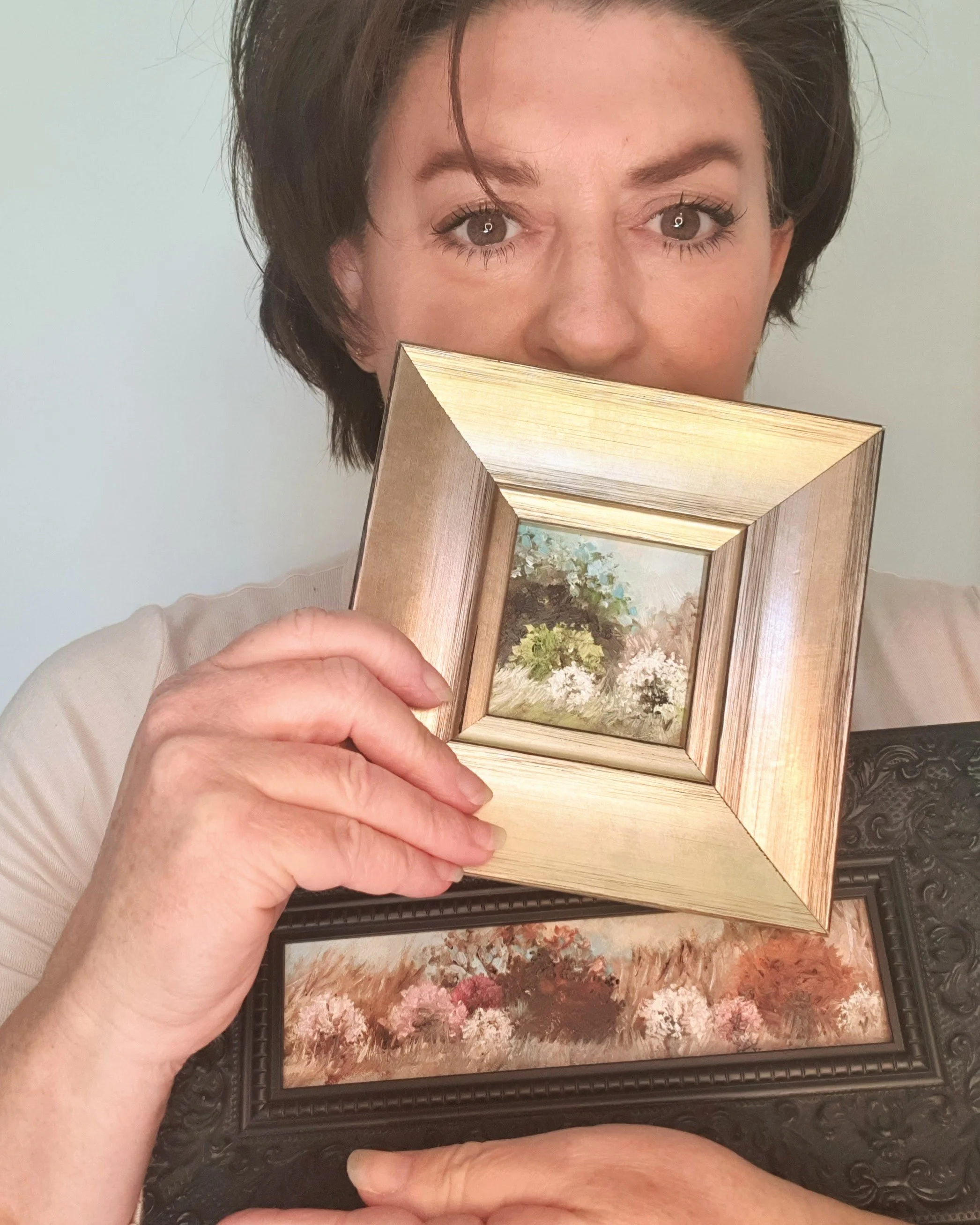 Australian artist Victoria Pynt holding framed miniature oil painting of rustic country scene with white blossom tree.
