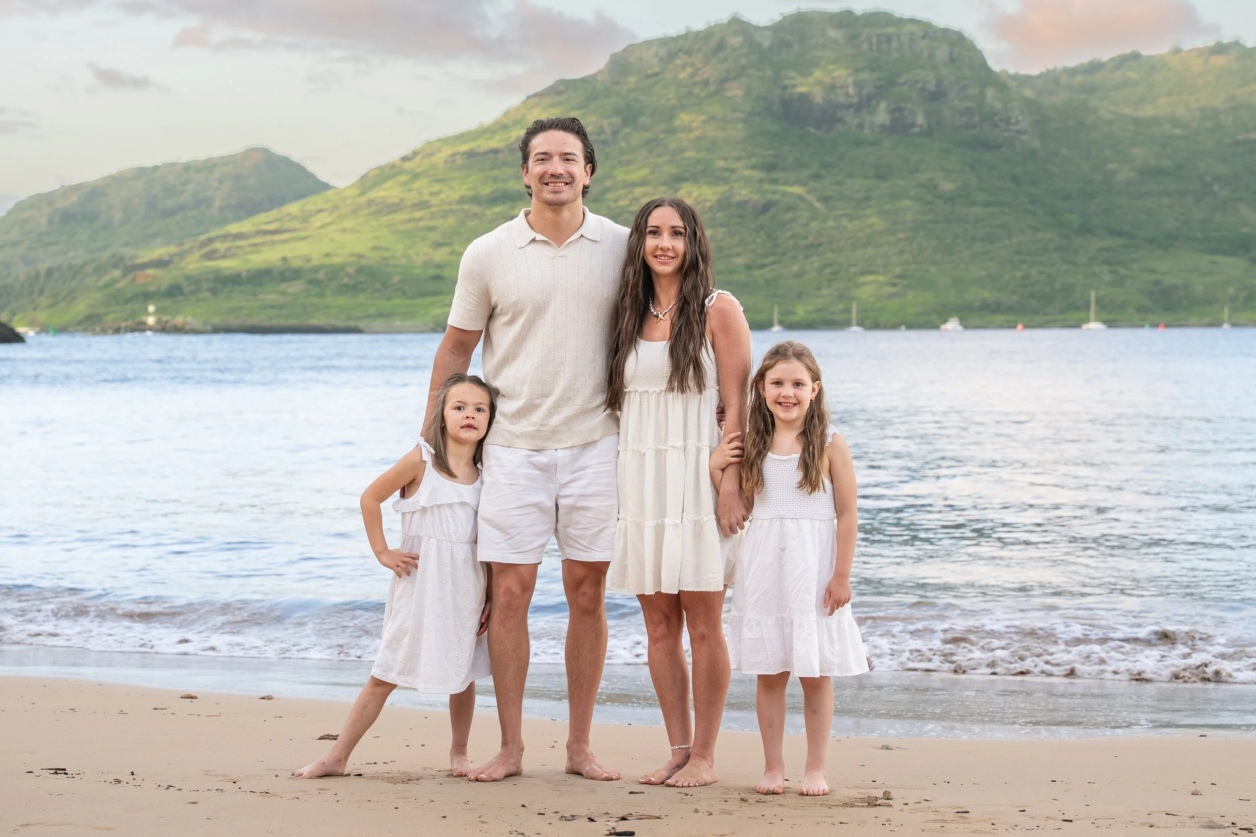 Family of four in white outfits posing on a Kauai beach with a mountain backdrop.