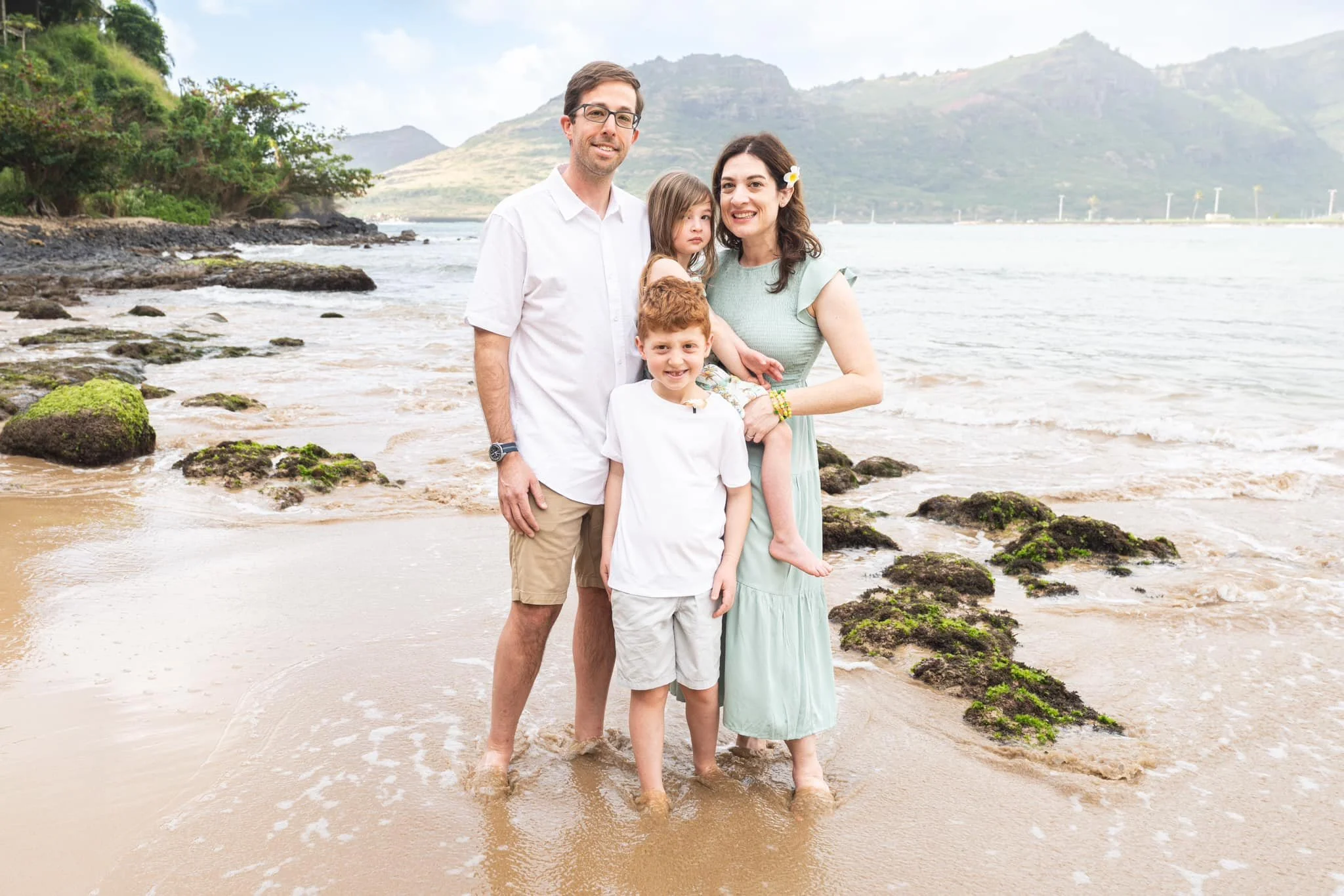 Family of four stands barefoot in gentle surf along a rocky Kauai coastline.