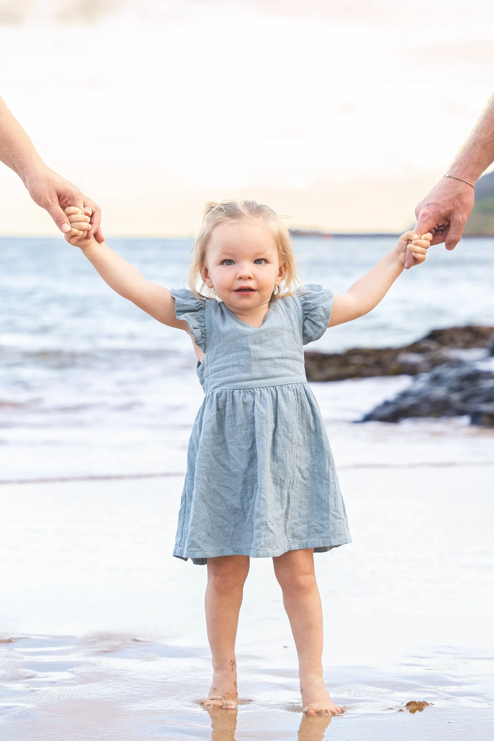 Toddler girl in a blue dress holding her parents' hands while standing on a Kauai beach.