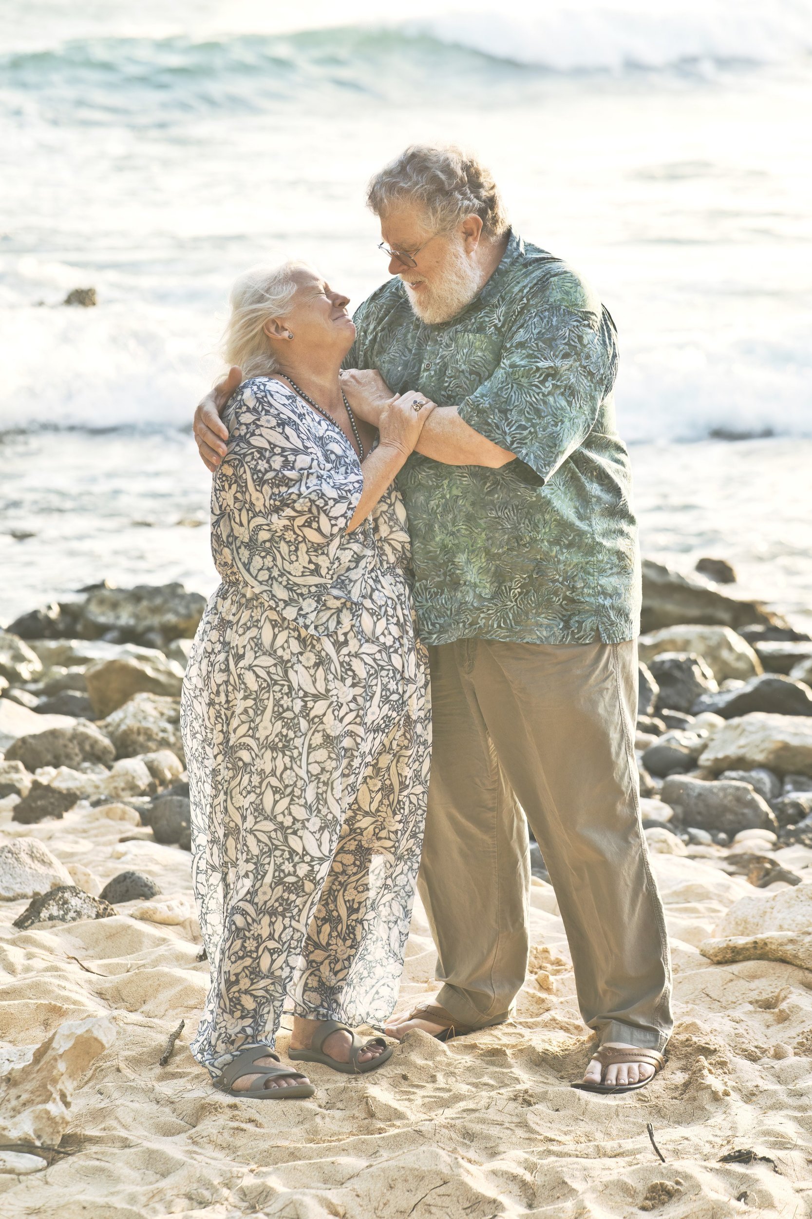 Grandparents laughing and dancing together on a sandy Kauai beach with waves behind them.