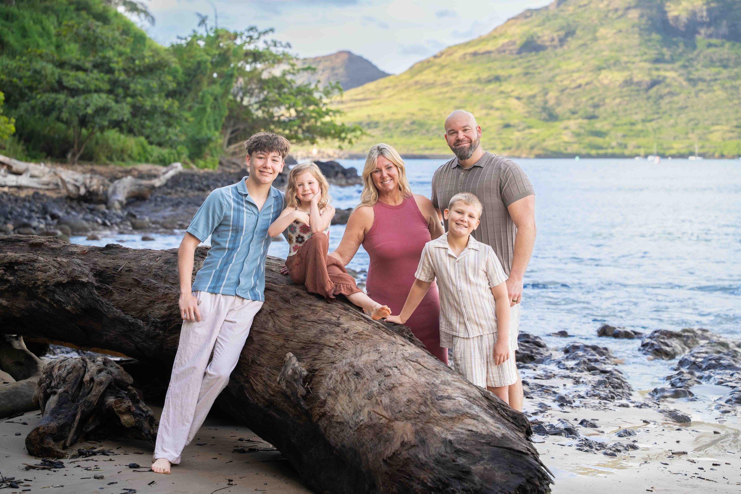 Family of five posing by a driftwood log on a rocky Kauai beach.