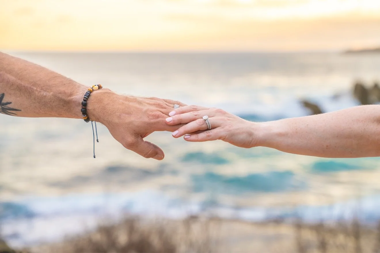 Close-up of a bride and groom's hands reaching for each other, their wedding rings sparkling with the ocean behind them on Kauai.