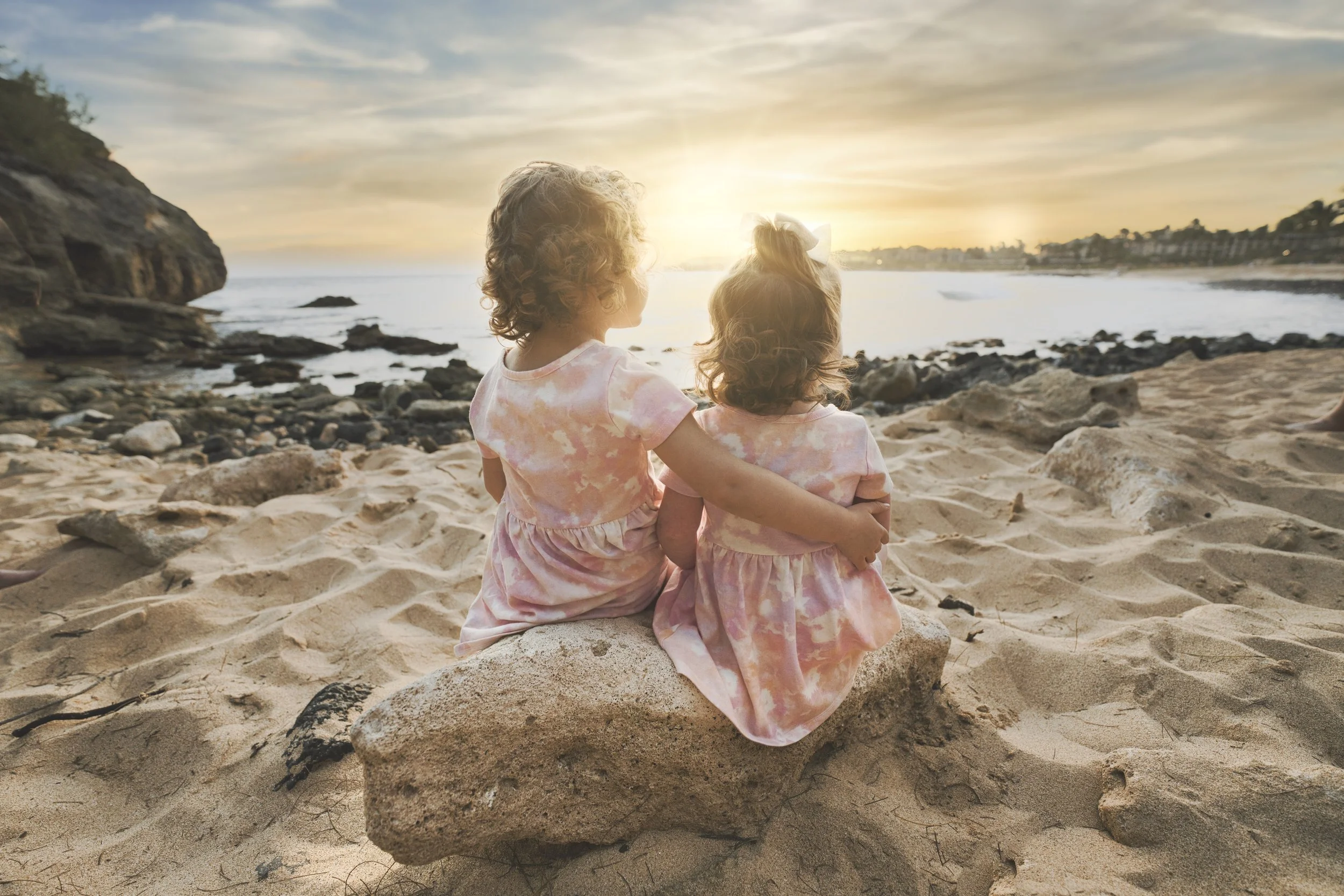 Two little sisters sitting on a rock watching the sunset over a Kauai beach.