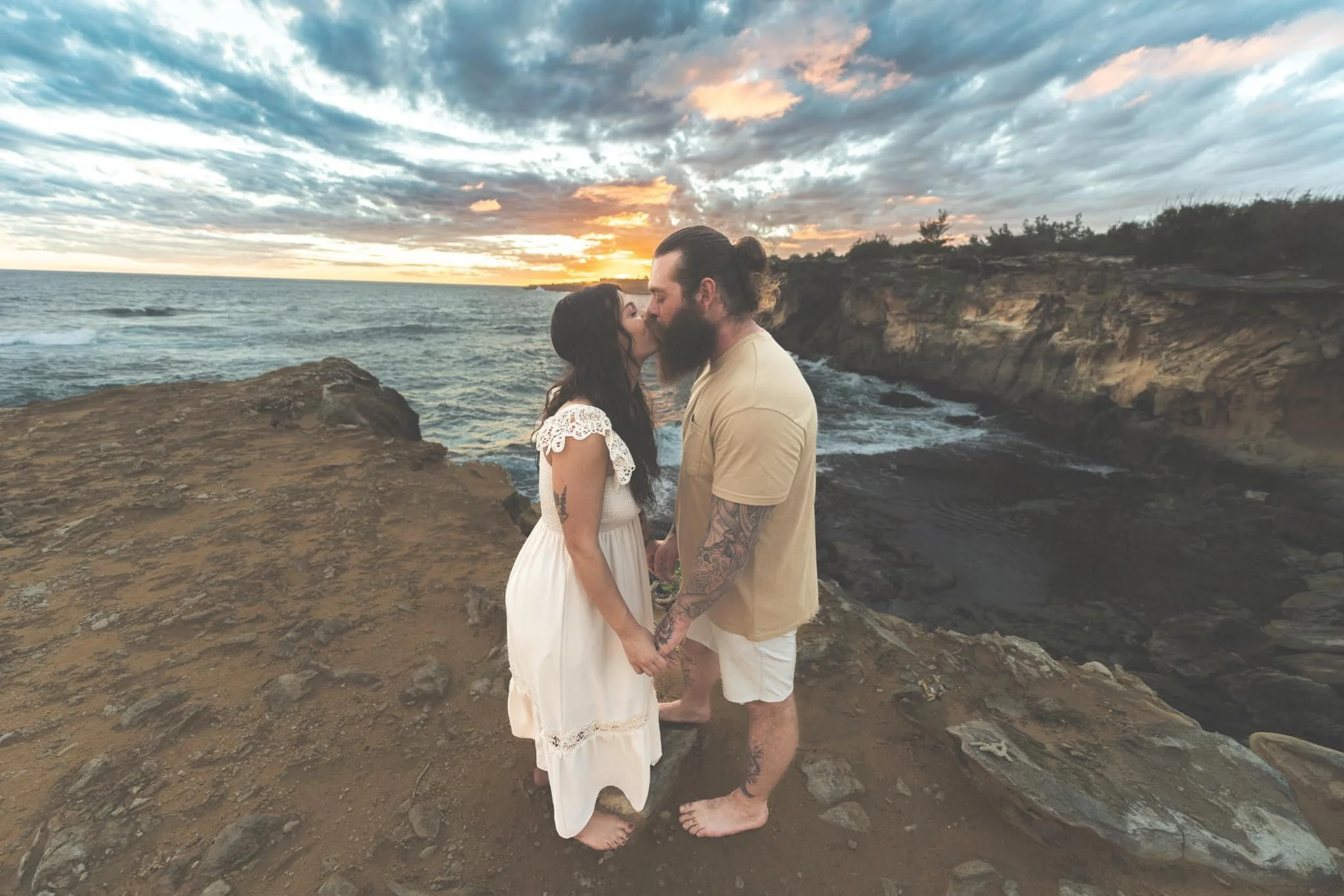 Couple kissing on a cliff edge under a dramatic sunset sky at Shipwreck Beach, Kauai.