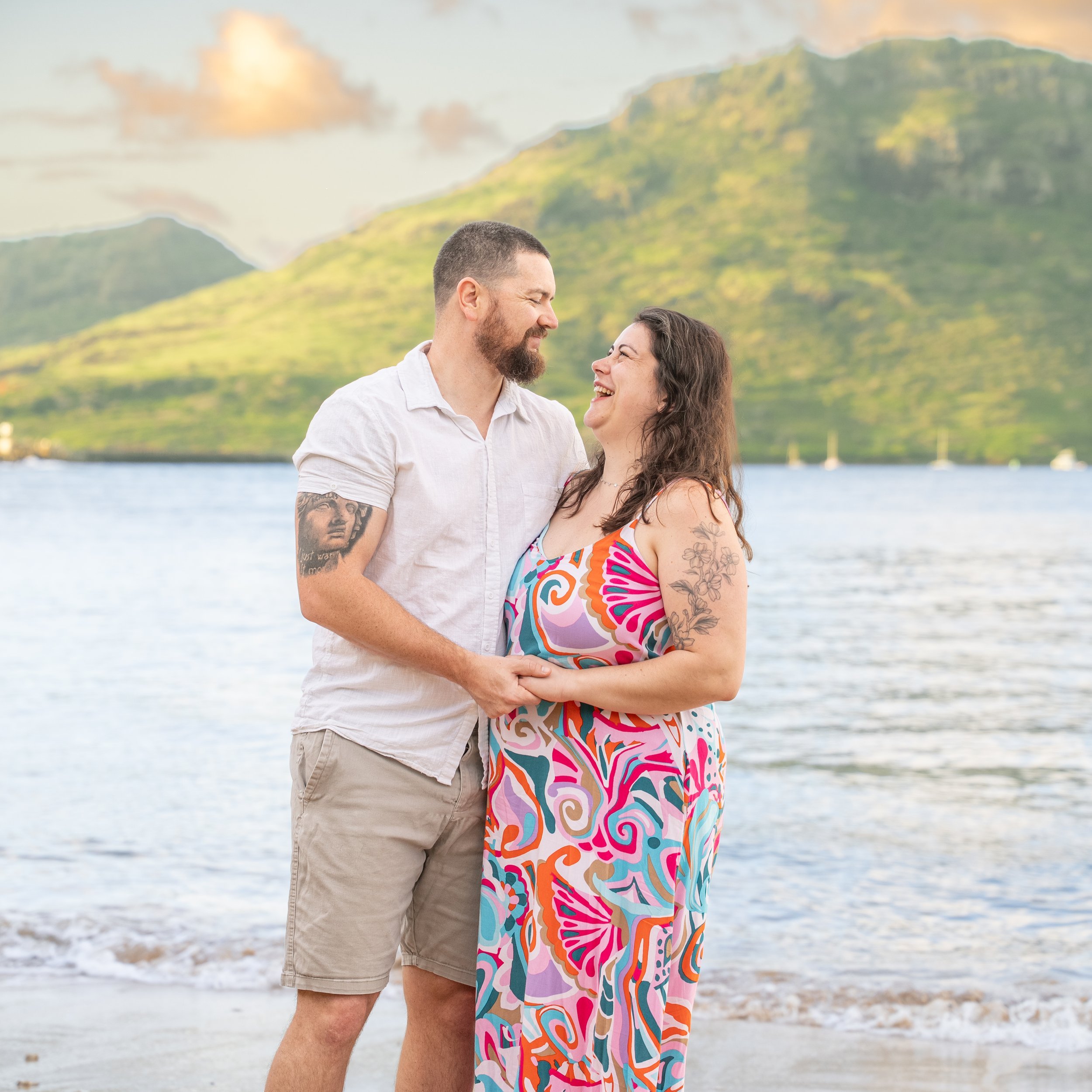 Laughing couple embracing on a Kauai beach with a bay and green mountains behind.
