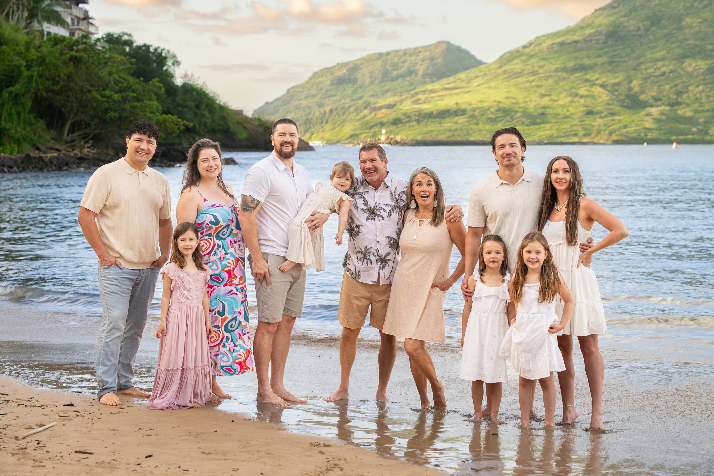 Large extended family posing at the water's edge on a Kauai beach with green mountains behind them.