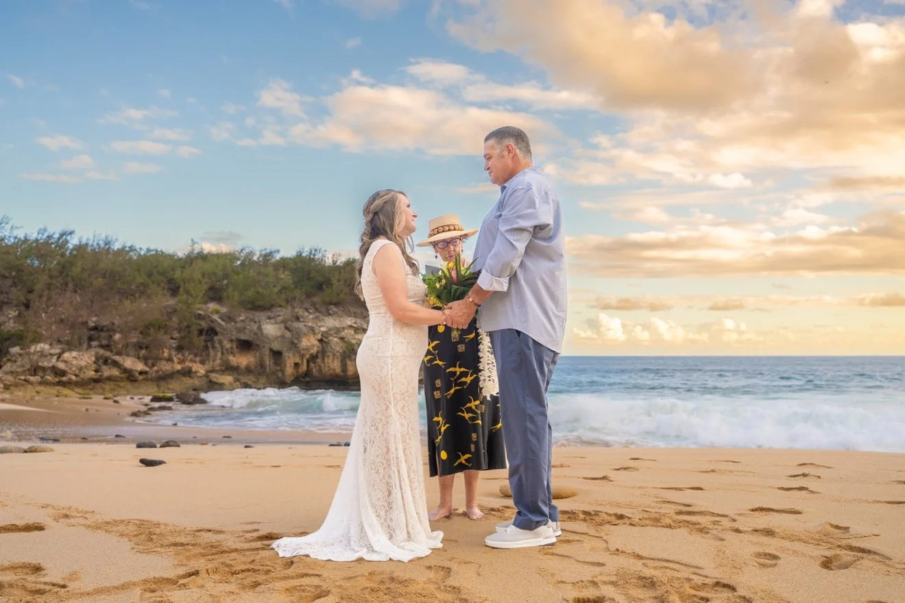 Bride and groom exchanging vows on a wide sandy Kauai beach with their officiant, waves rolling in behind them under a golden sky.