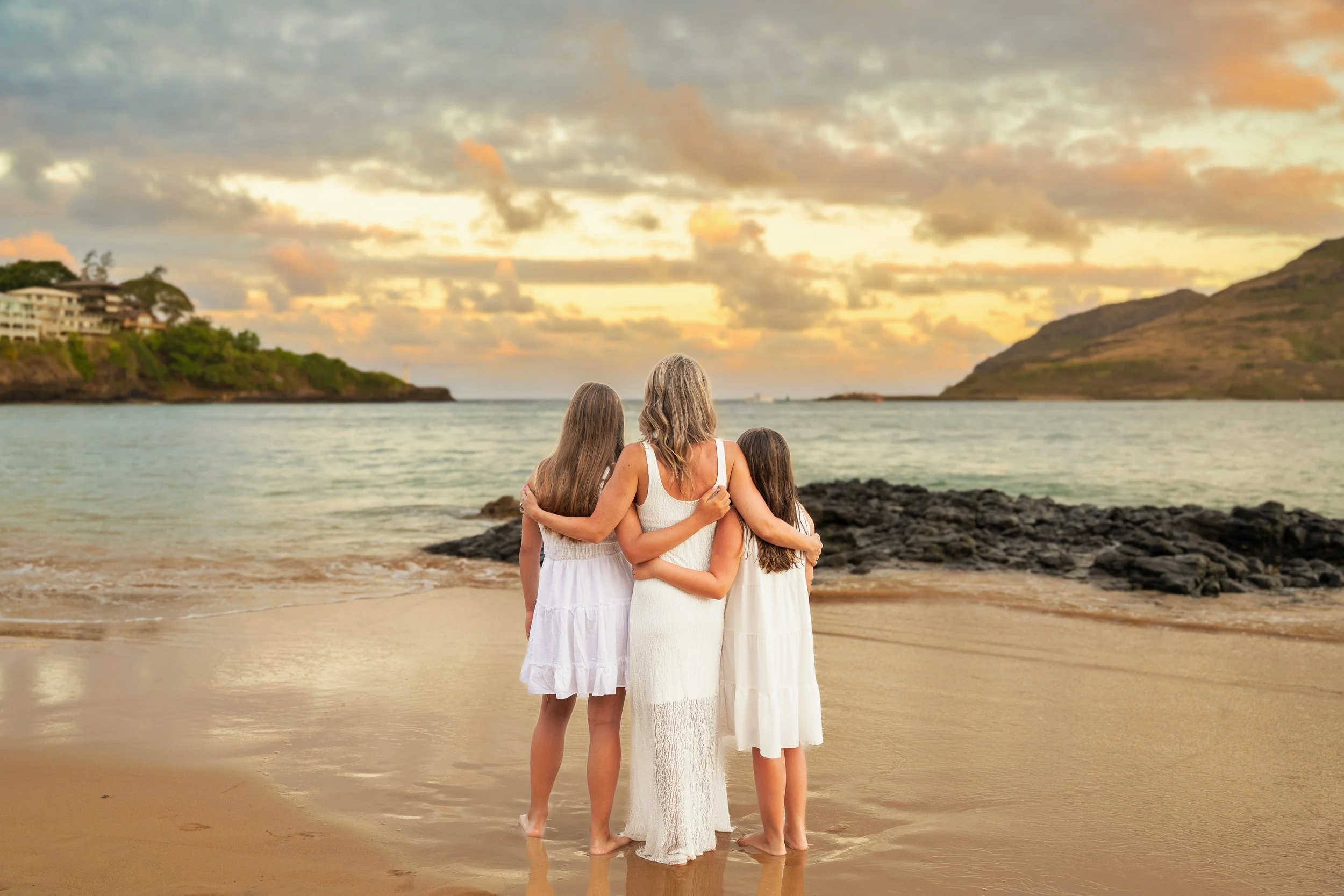 Mom with her arms around her daughters watching the sunset over the ocean in Kauai.