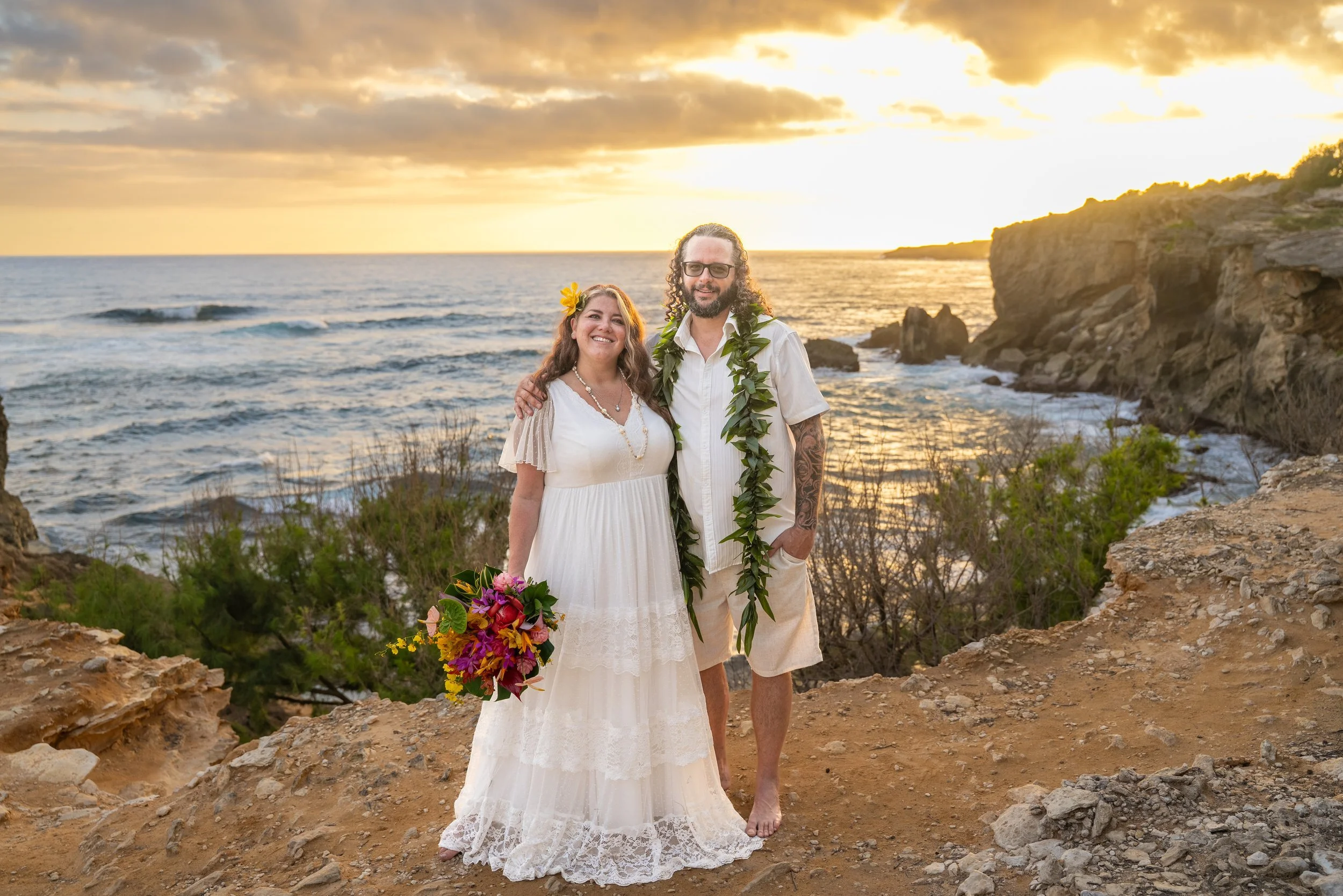 Newlywed couple posing on a rocky ocean cliff in Kauai at sunset, the bride holding a tropical bouquet.