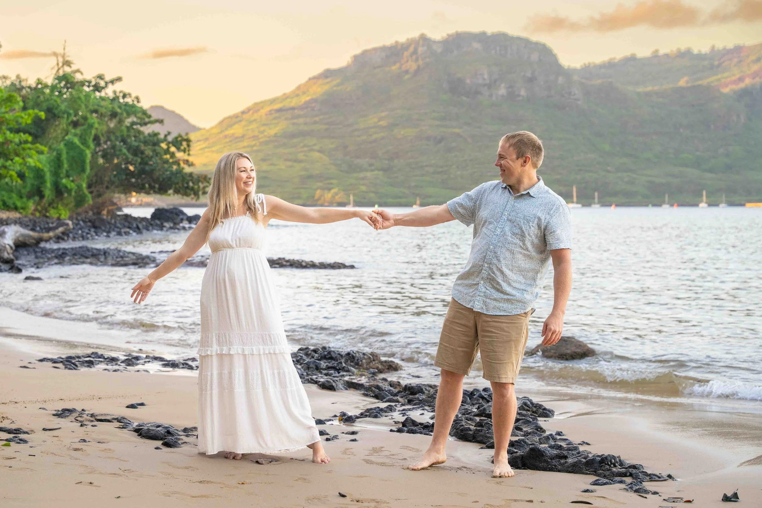 Playful couple holding hands on a Kauai beach with a mountain behind them.