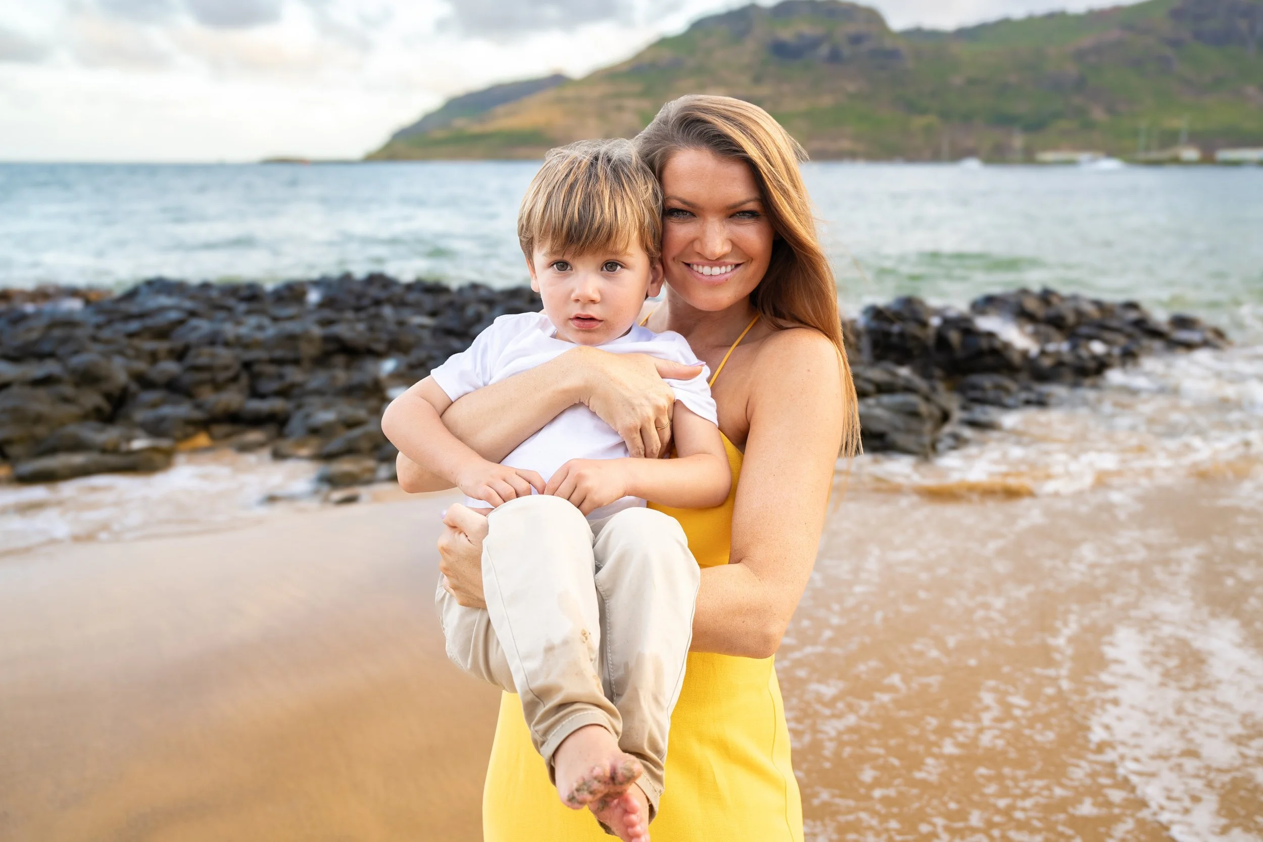 Mom in a yellow dress holding her young son on a rocky Kauai beach.