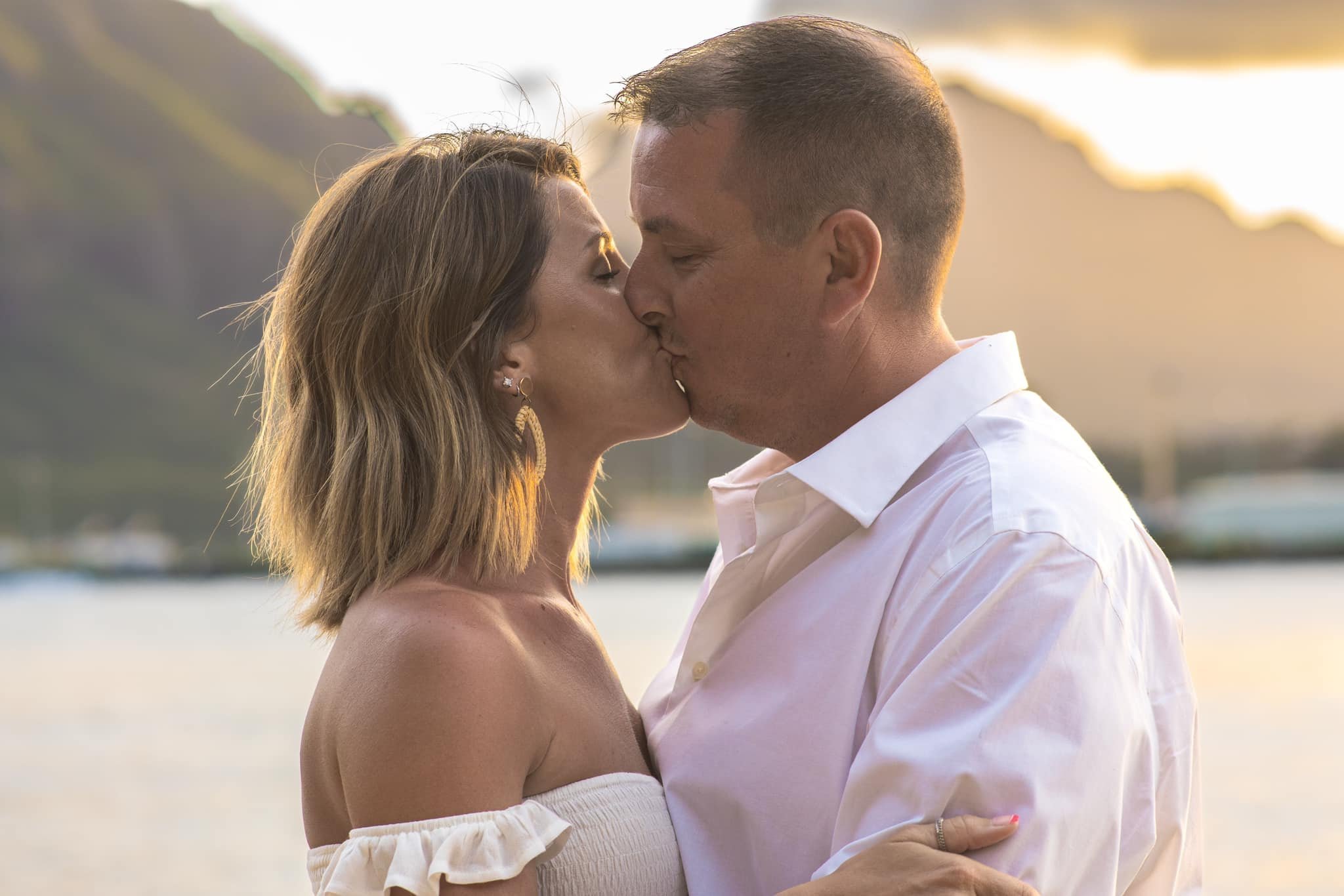 Couple kissing at sunset on a Kauai beach with mountains behind them.