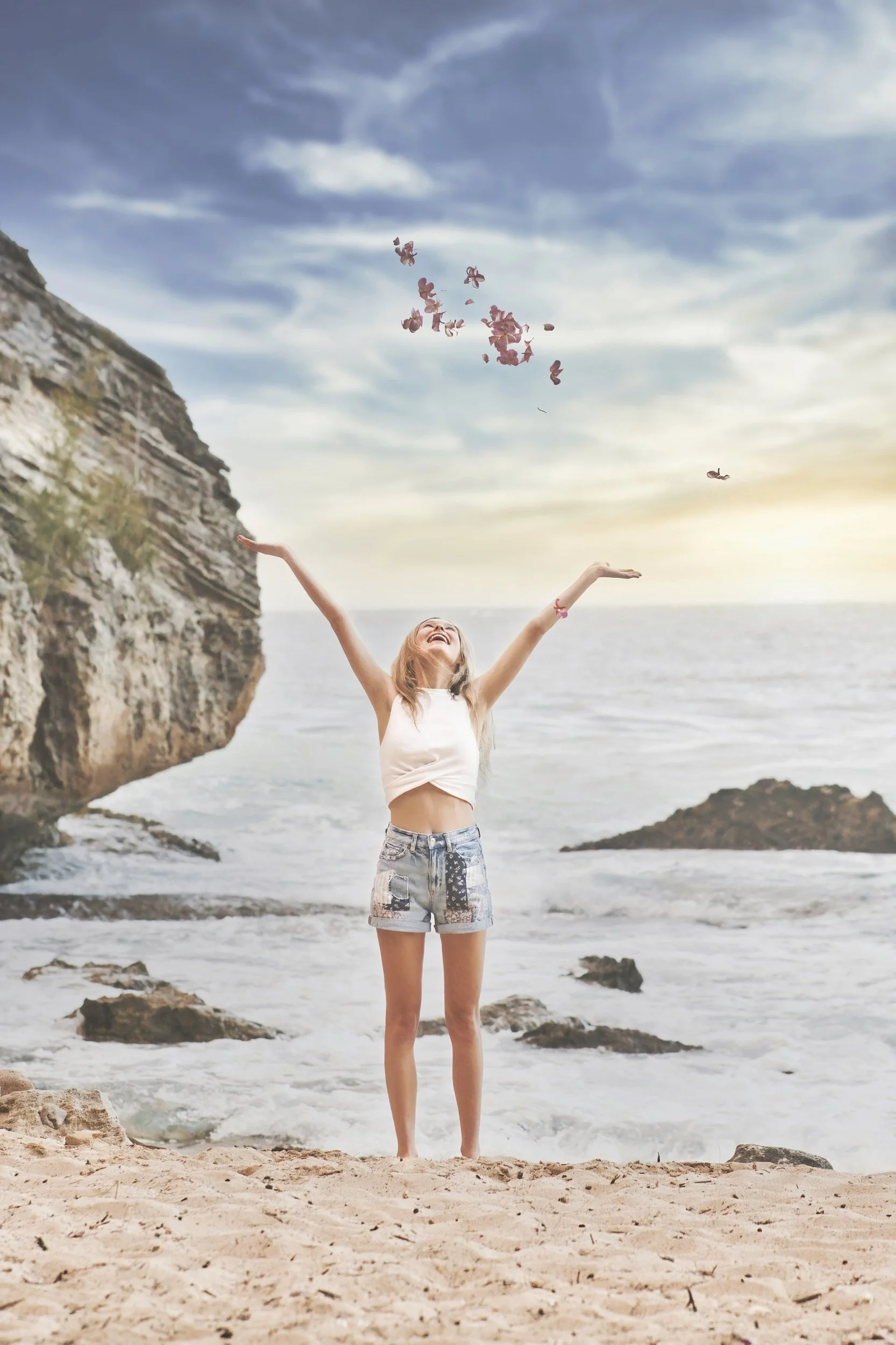 Woman tossing flower petals with arms raised on the beach at Shipwreck Beach, Kauai.