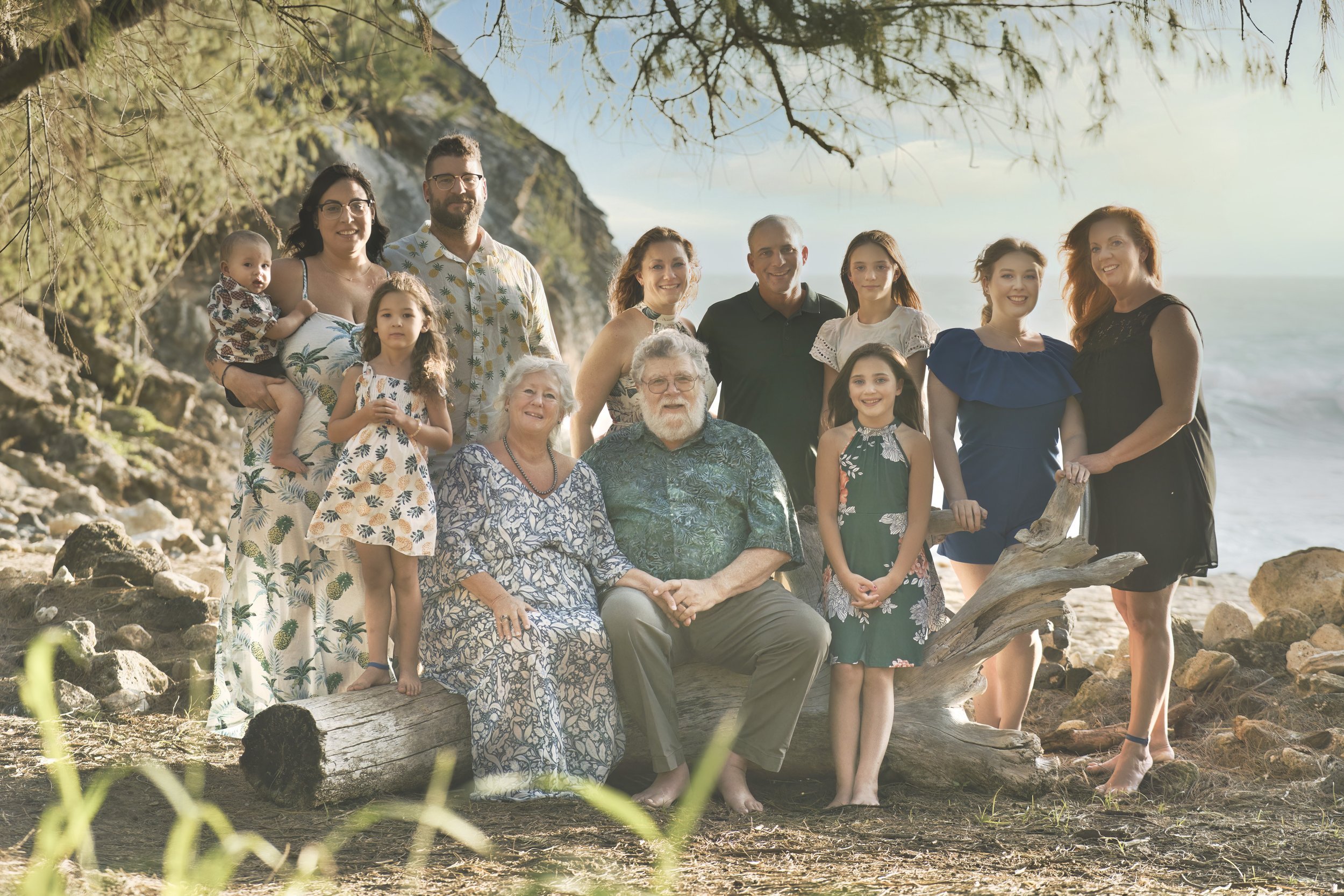 Large extended family gathered around driftwood on a Kauai beach with grandparents seated in front.