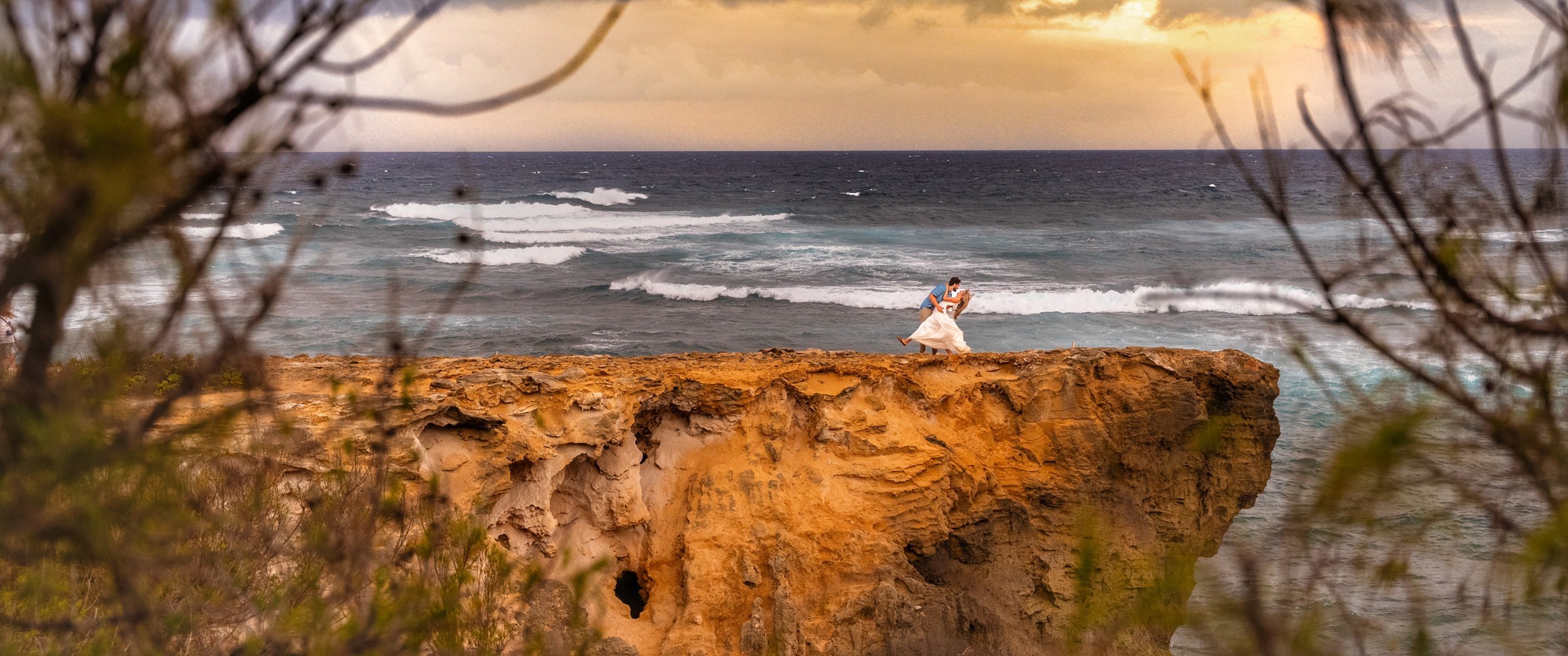 Couple sitting on the edge of a tall sea cliff overlooking the ocean in Kauai.