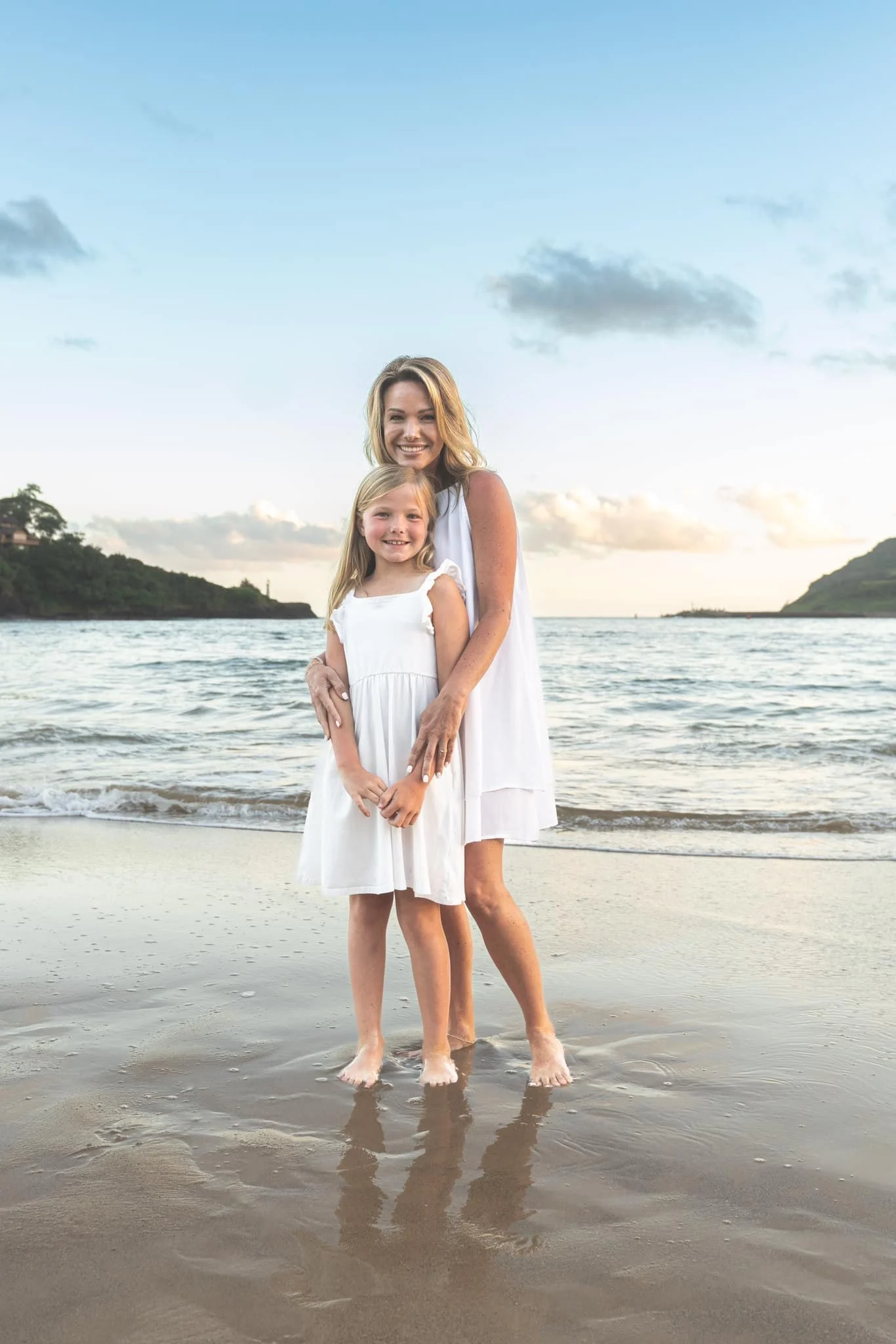 Mom standing behind her daughter, both in white dresses, in the surf on Kauai.