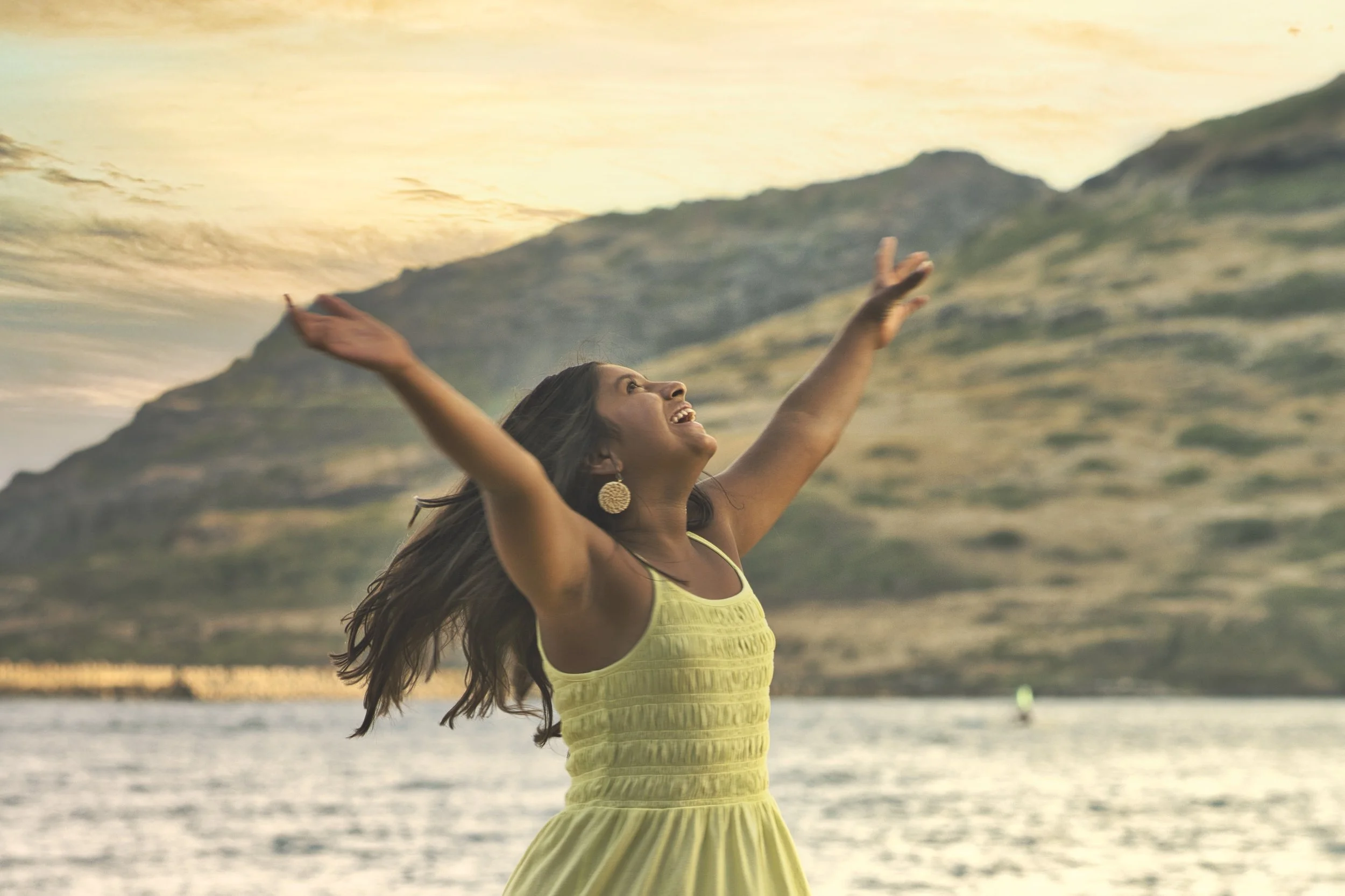 Young girl in a yellow dress laughing with arms raised on a Kauai beach.