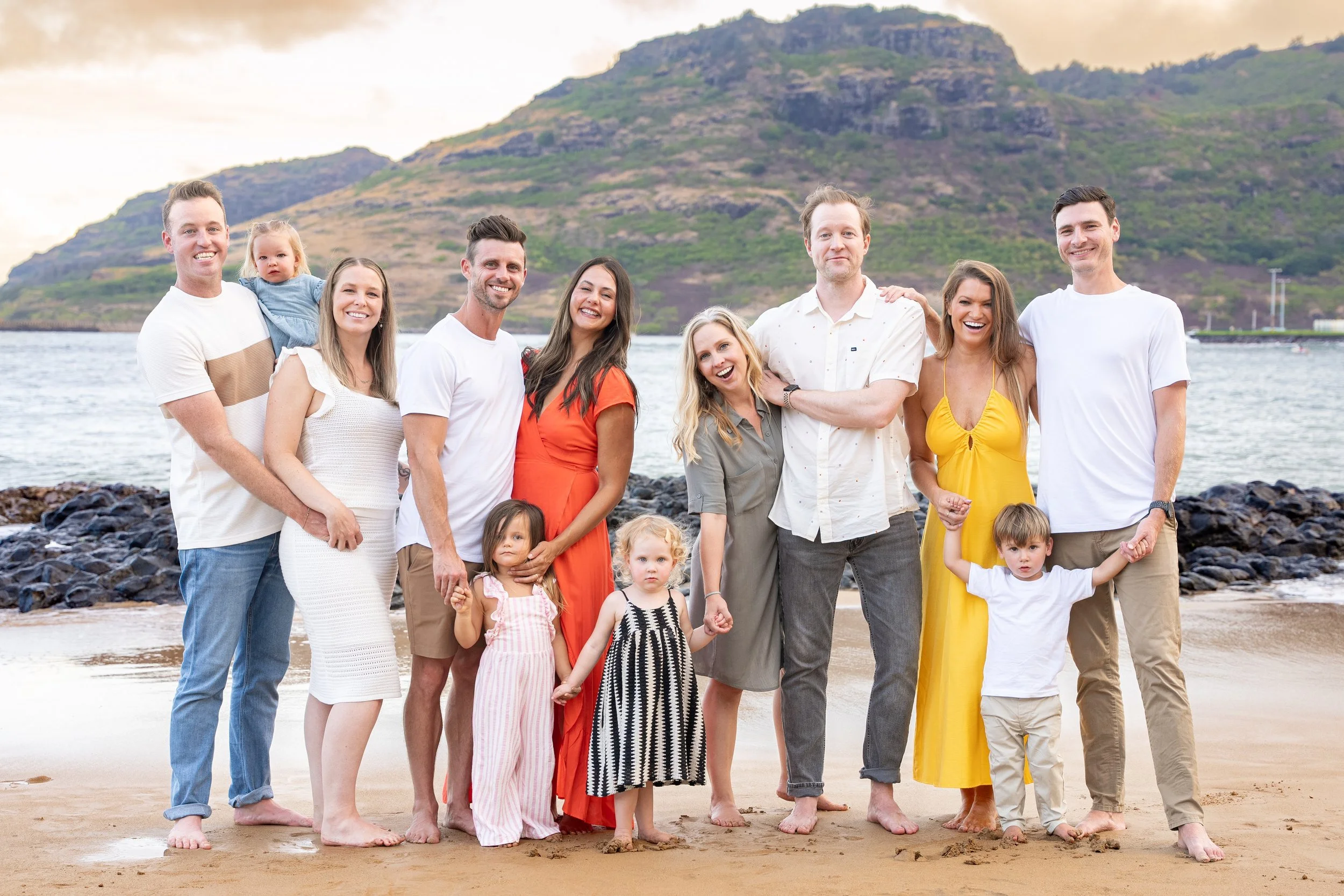 Large multi-generational family posing together on a Kauai beach with mountains behind them.