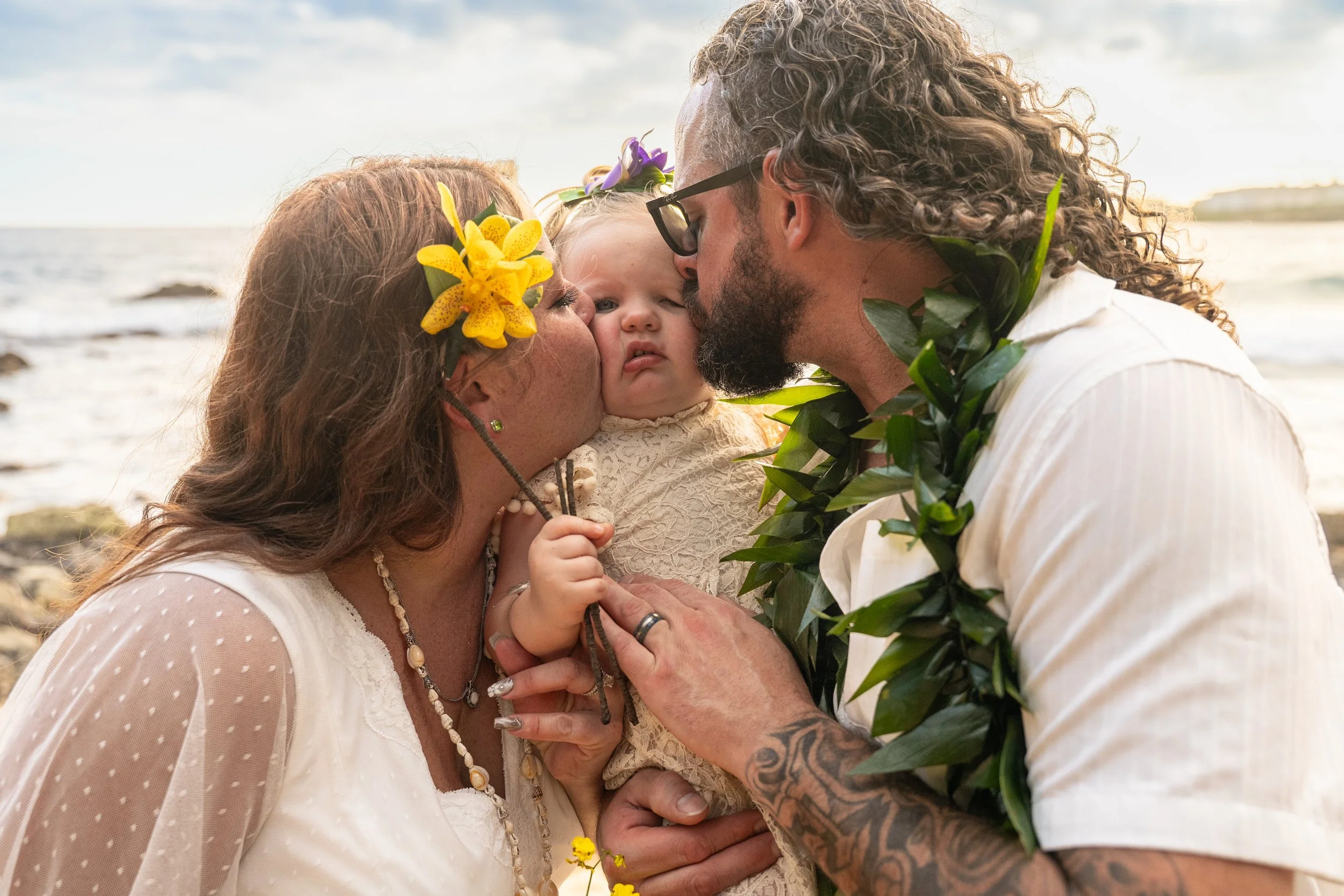 Newlywed couple kissing their baby's cheeks on a Kauai beach.