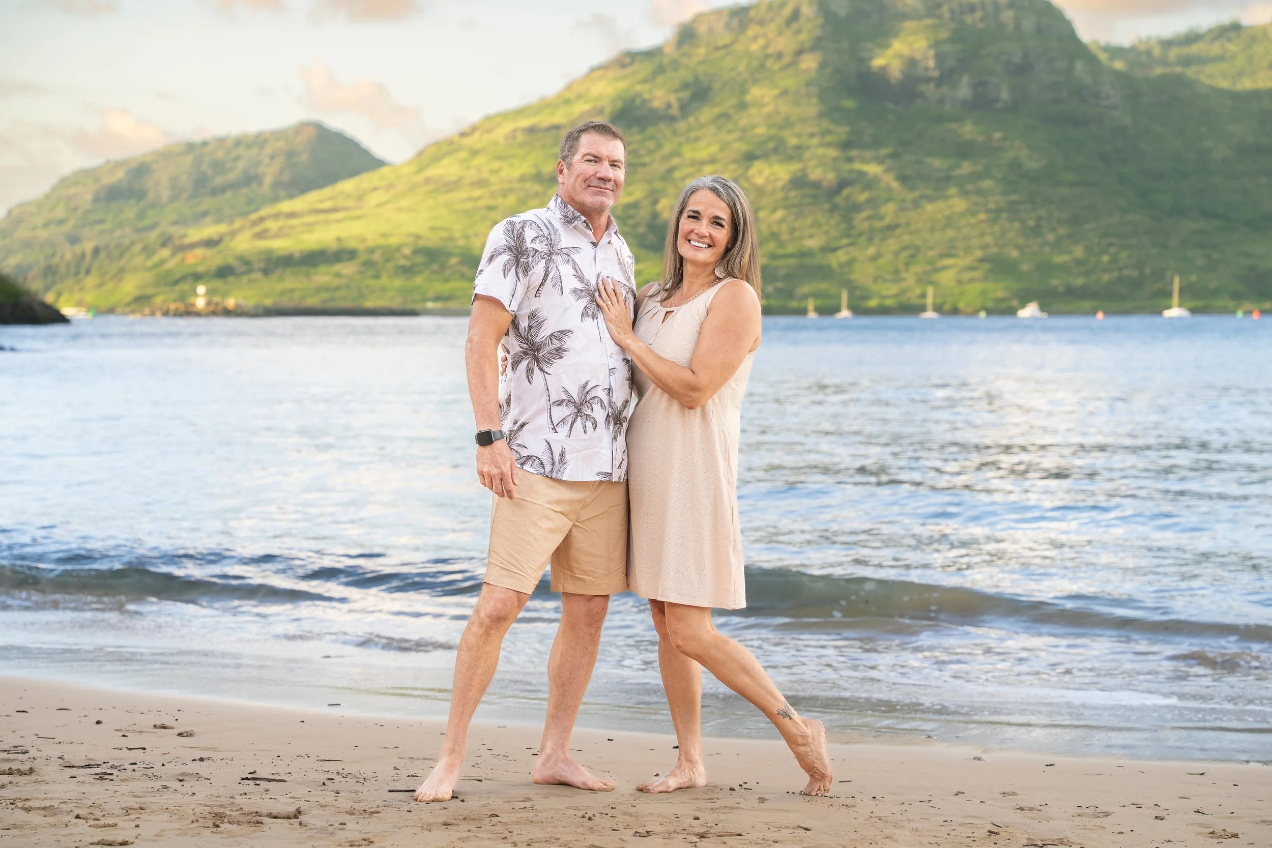 Smiling couple posing together on a Kauai beach with lush mountains behind them.