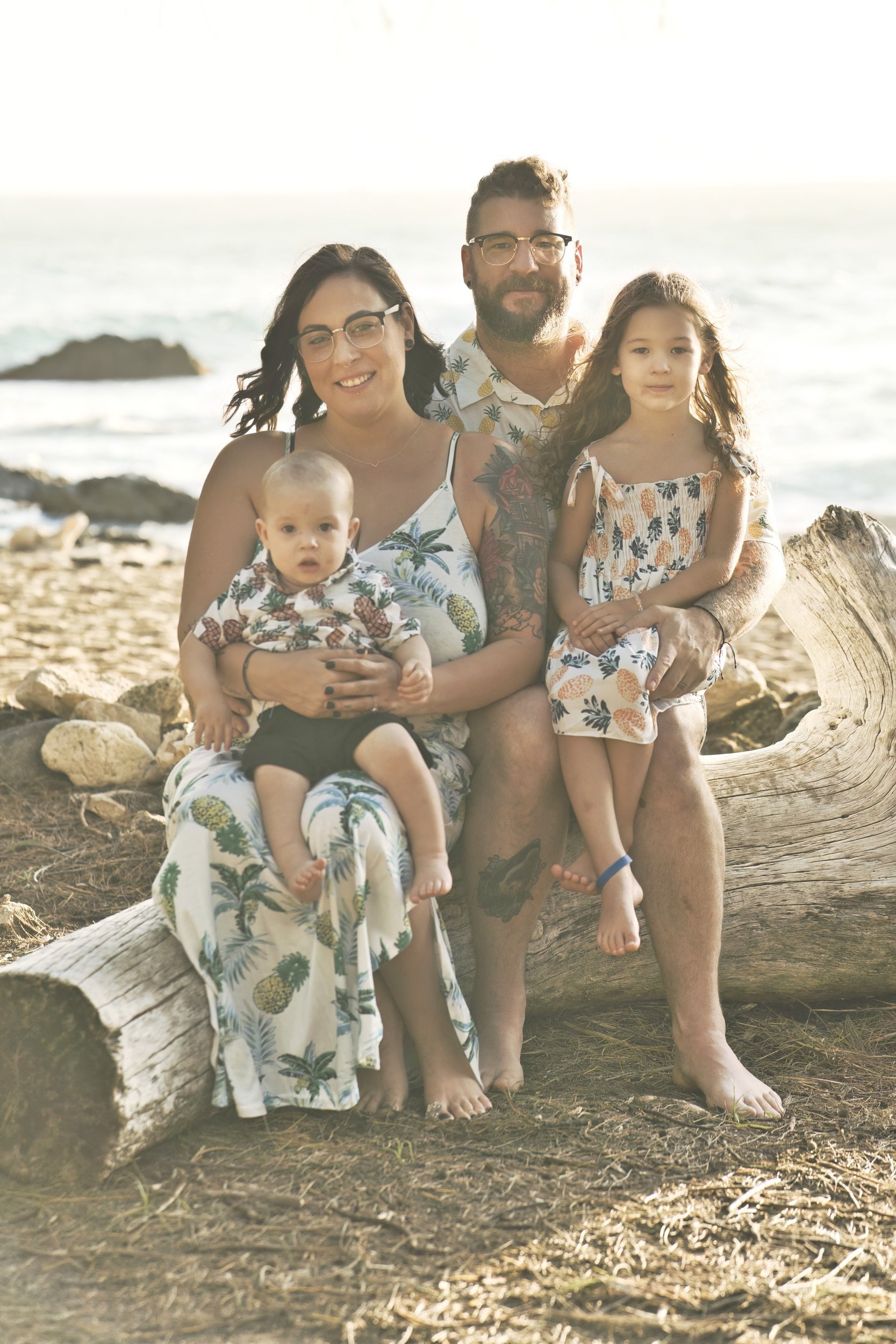 Family of four in pineapple-print outfits sitting on driftwood at a rocky Kauai beach.