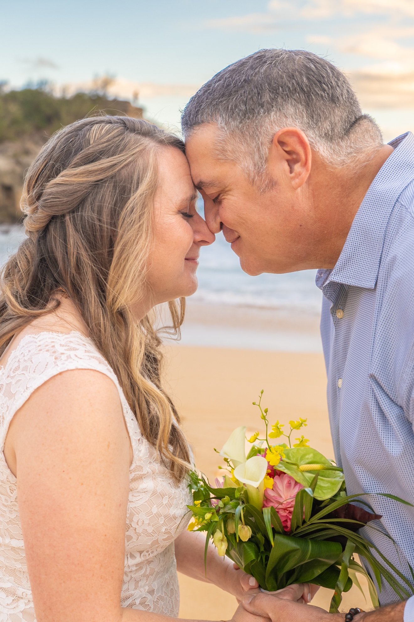 Bride and groom touching foreheads with their eyes closed, holding a colorful tropical bouquet together on a Kauai beach.