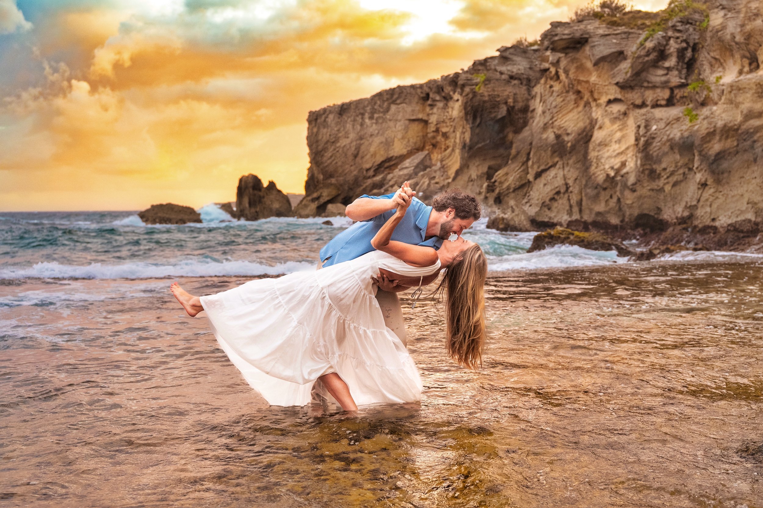 Groom dipping his bride for a kiss in the shallow water beside rocky cliffs on Kauai at sunset.