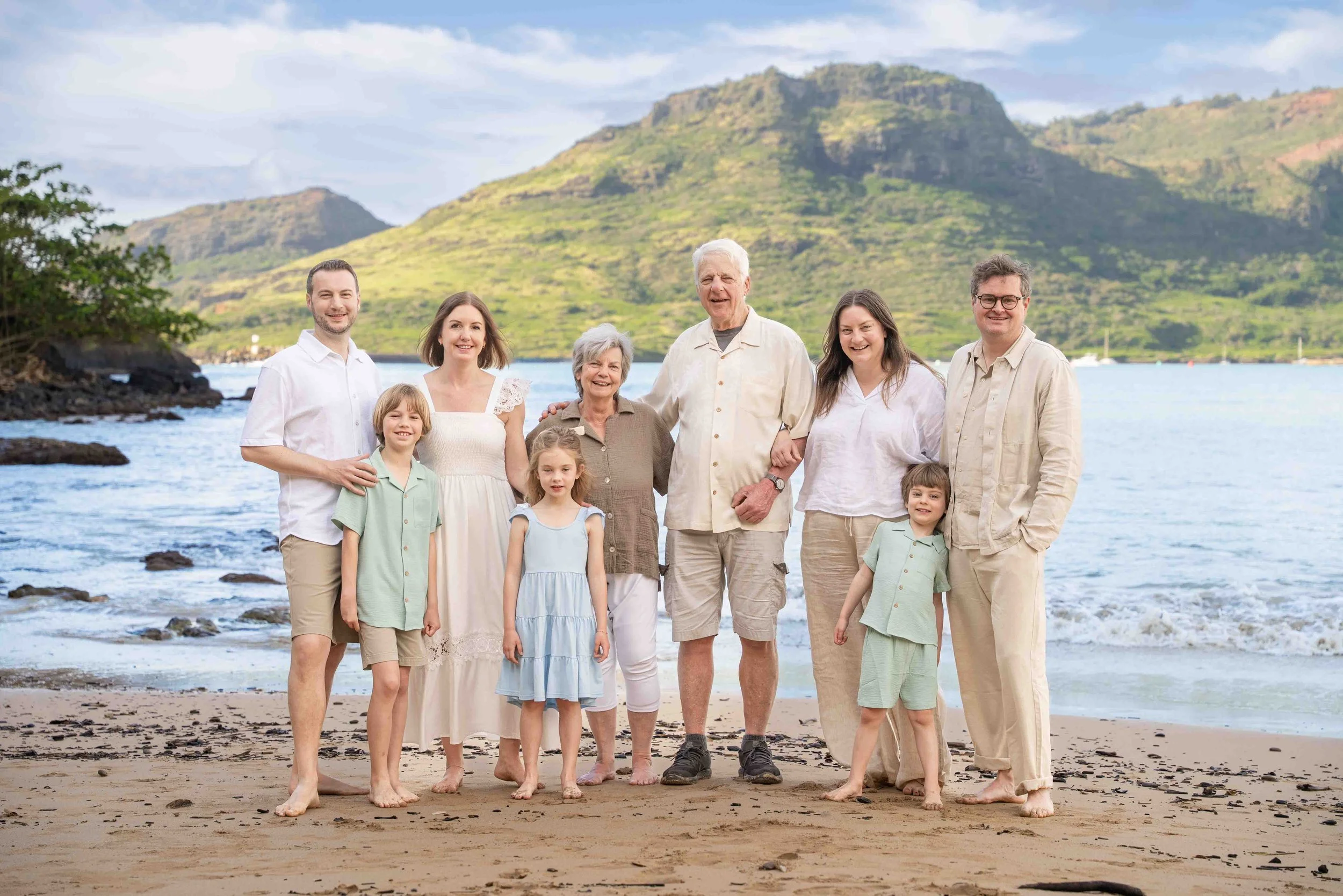 Nine-person extended family smiles together on a Kauai beach with green mountains across the bay.