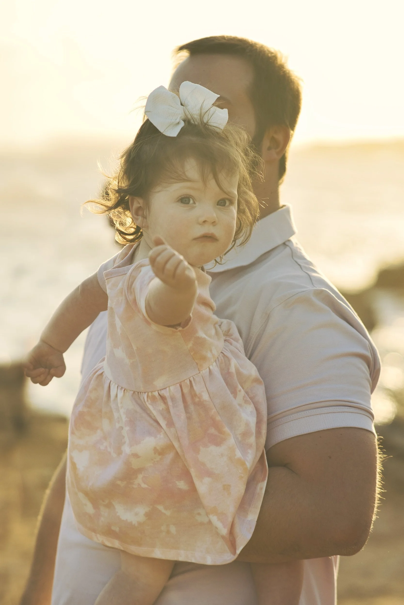 Dad holding his toddler daughter with a big bow as she points at the camera on a Kauai beach at sunset.