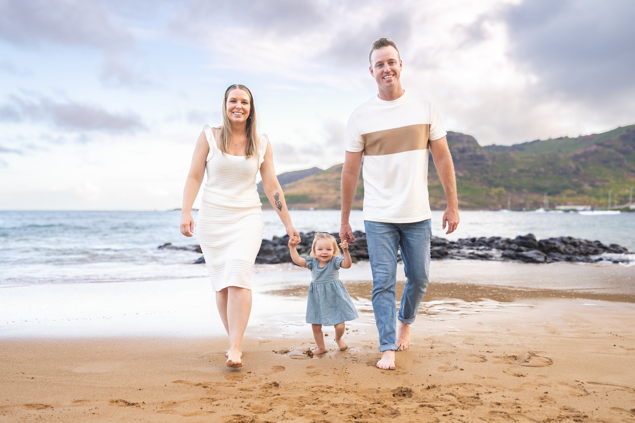 Family of three walking hand in hand toward the camera on a sandy Kauai beach.