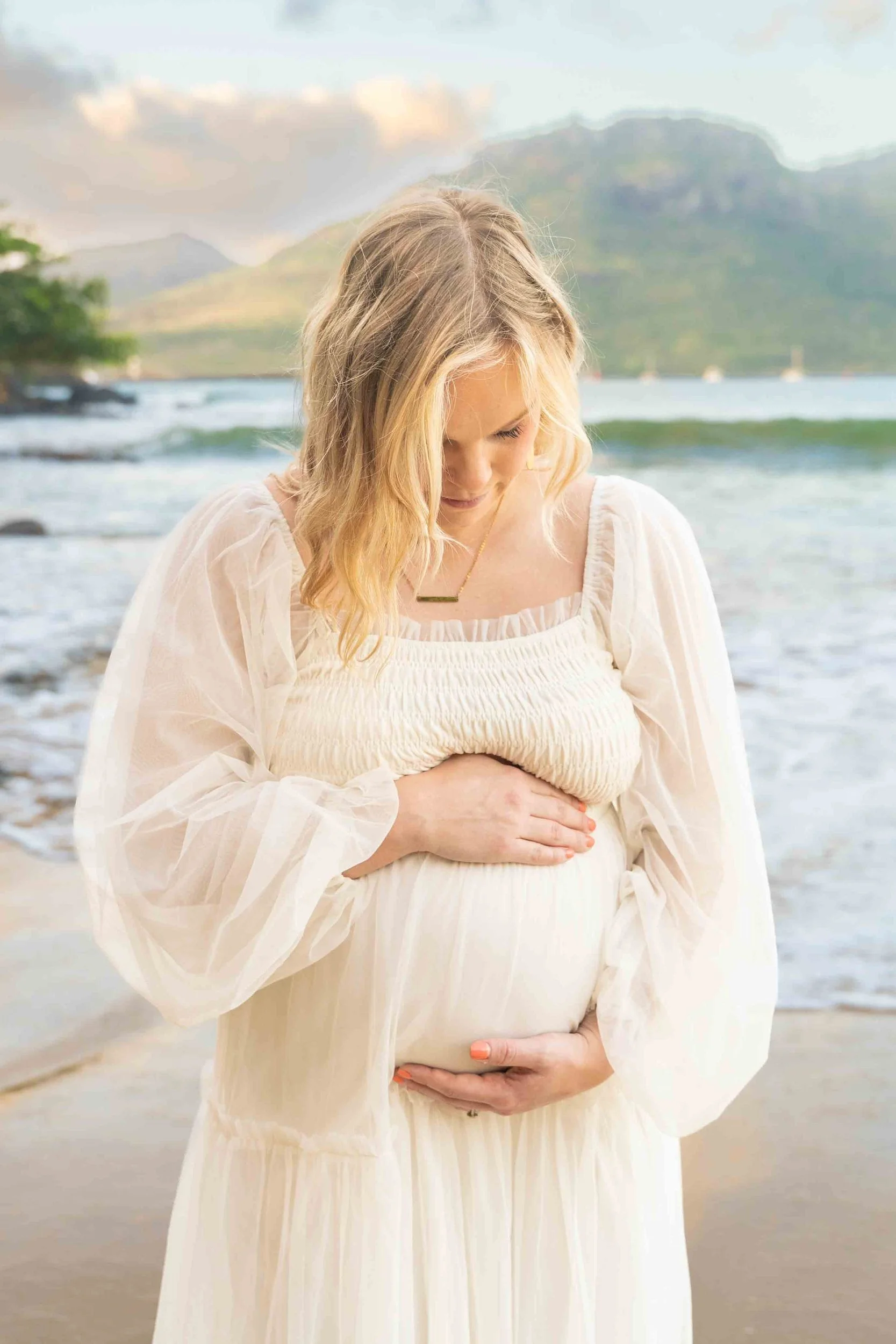 Expecting mom in a white dress looking down at her belly on a Kauai beach.