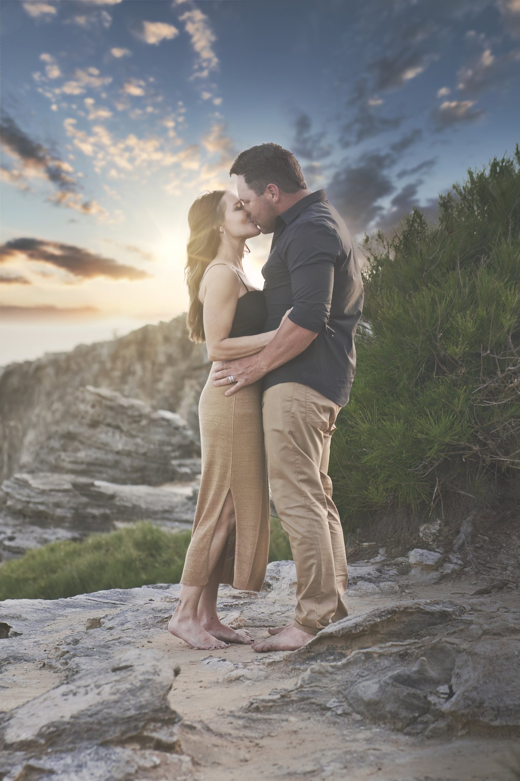Couple sharing a kiss on a rocky cliff top at Shipwreck Beach, Kauai, at sunset.