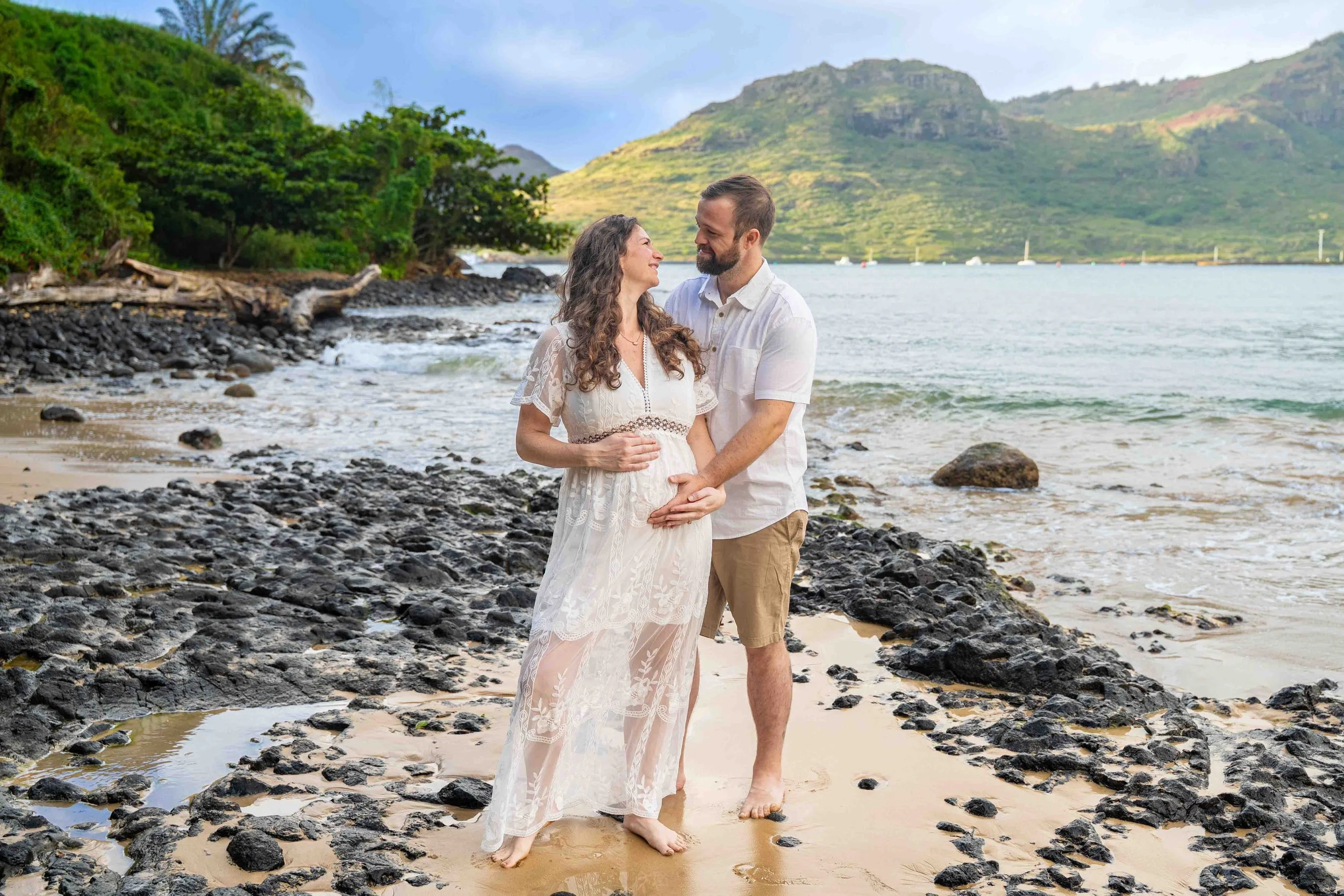 Man kissing his pregnant partner's head on a rocky Kauai shore.