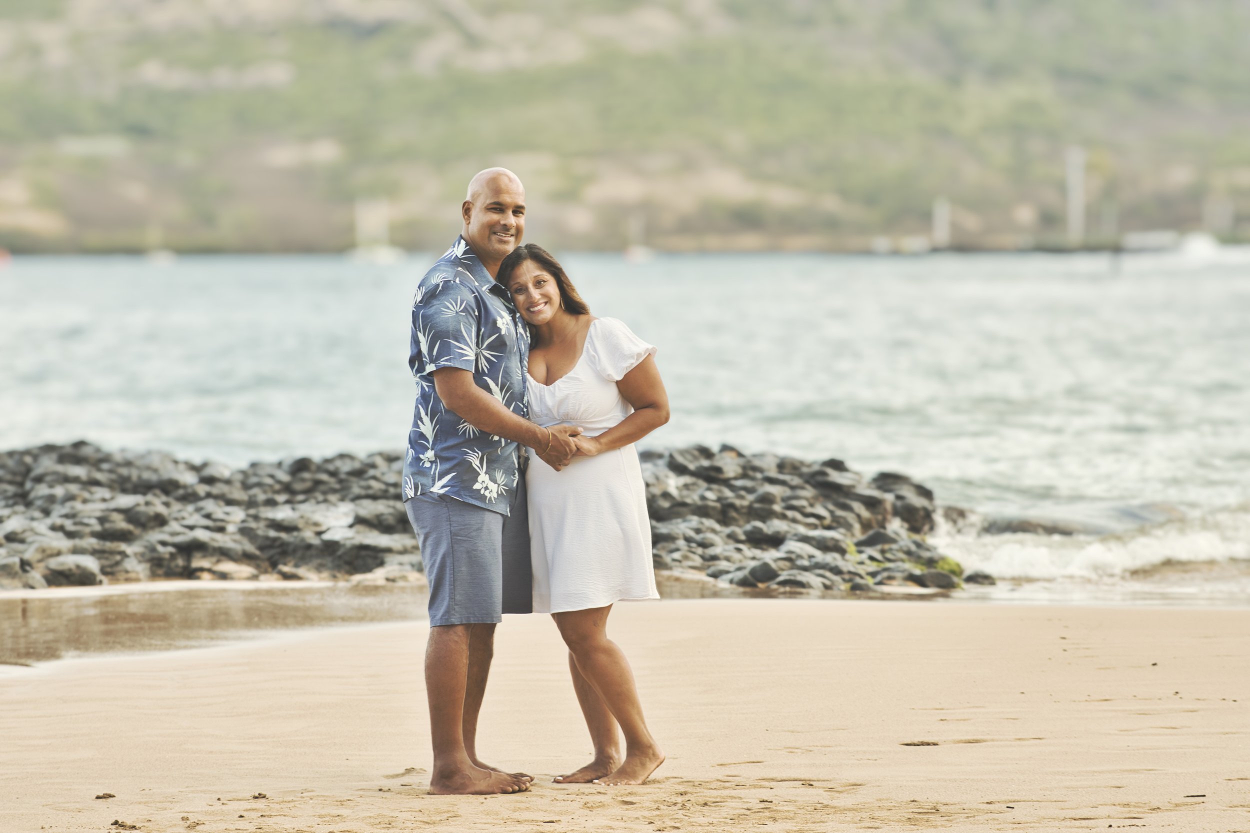 Couple embracing on a Kauai beach with a calm bay and rocky shoreline behind them.