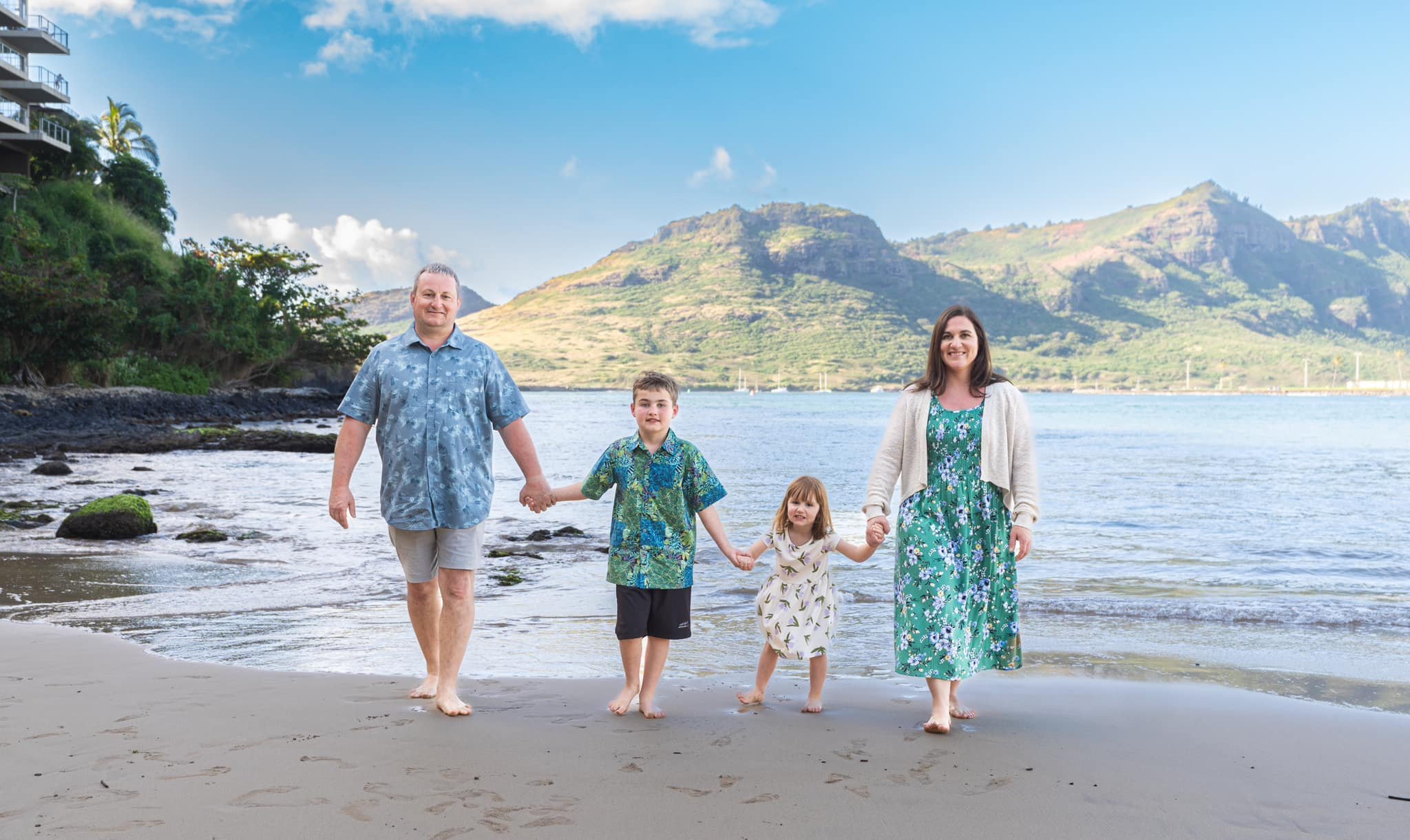 Family of four holding hands at the water's edge of a Kauai beach beneath green mountains.