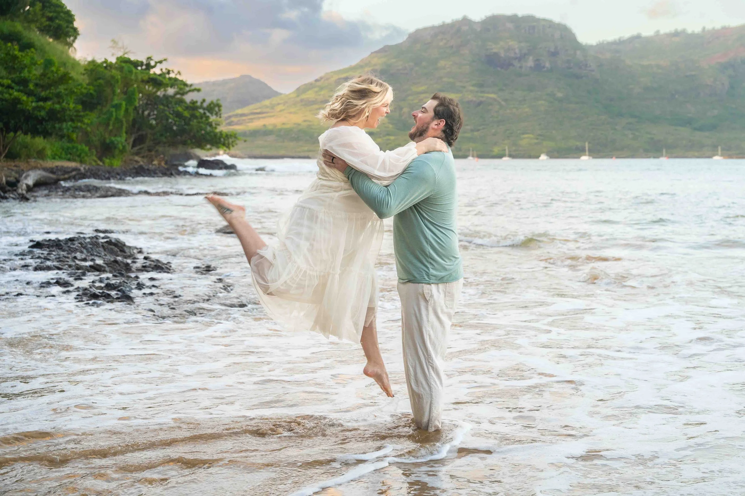 Man lifting his pregnant partner as they laugh in the surf on a Kauai beach.