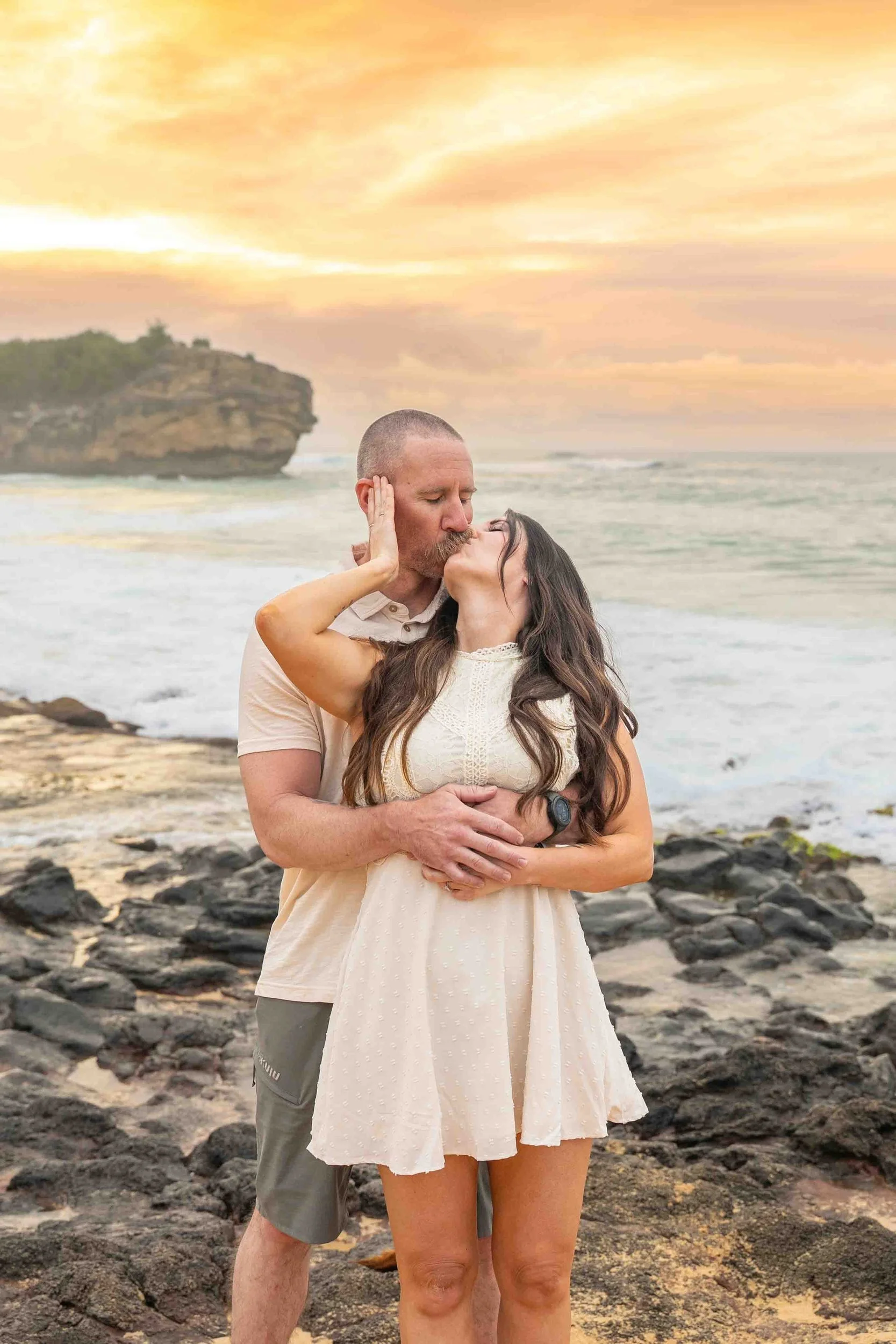 Couple embracing on the rocky shore at Shipwreck Beach, Kauai, at sunset.