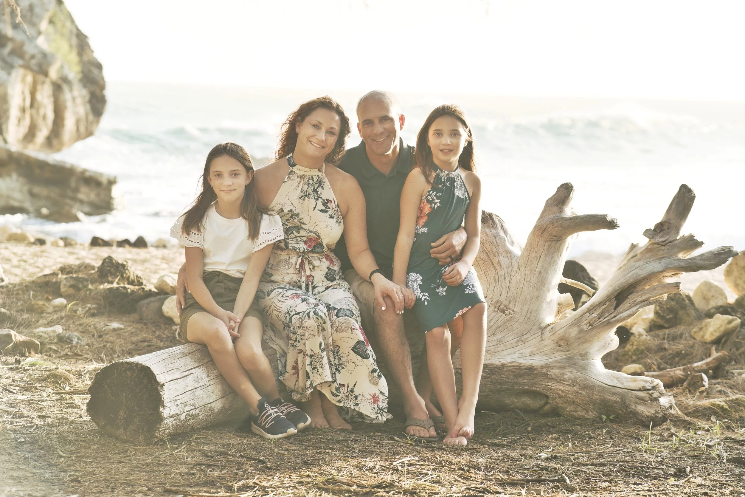 Family of four sitting together on driftwood at a rocky Kauai beach.