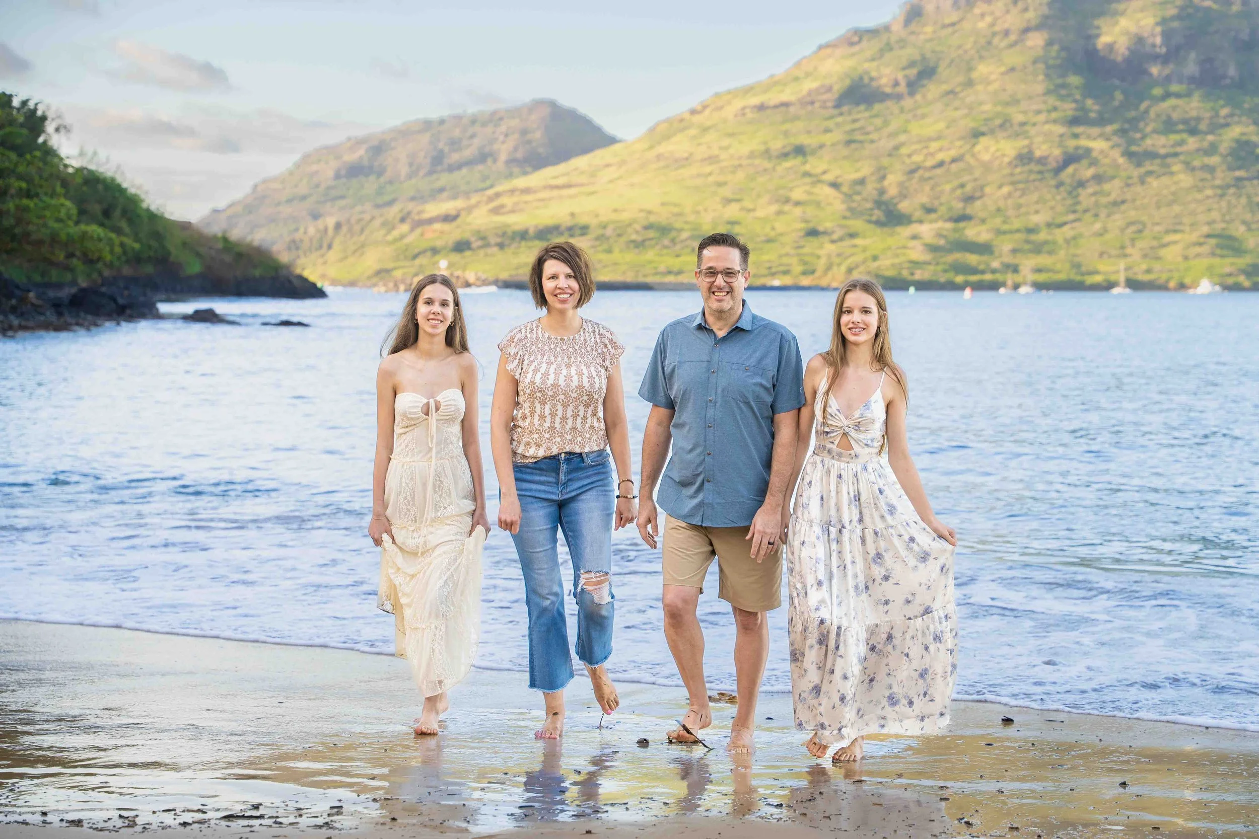 Family with two teenage daughters walking on a Kauai beach with mountains behind them.