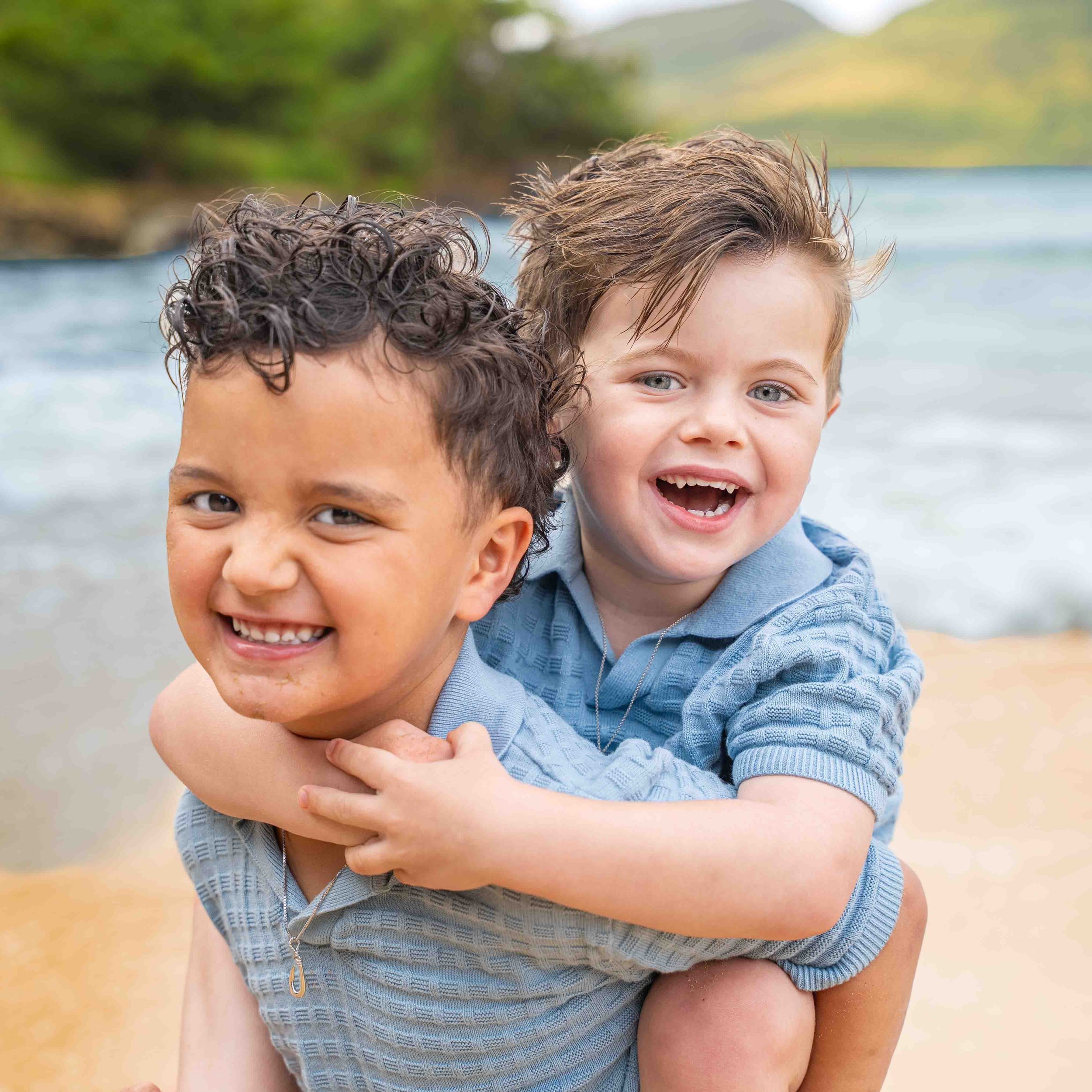 Two young boys laughing together in a piggyback hug on a Kauai beach.