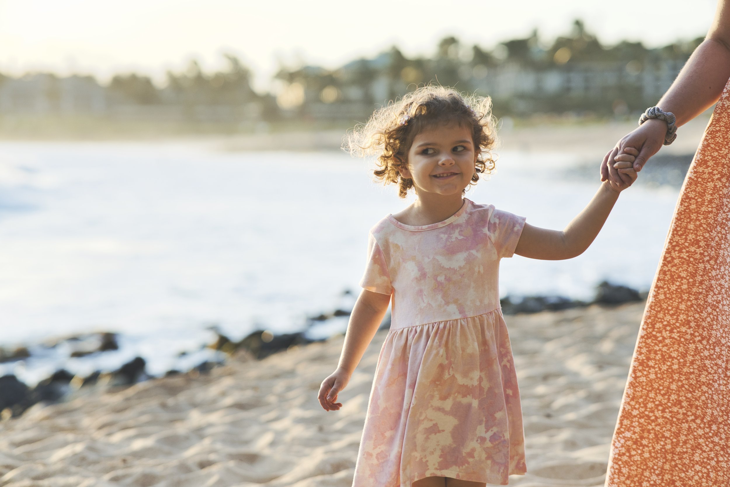Little girl in a pink dress holding her mom's hand on a Kauai beach at golden hour.