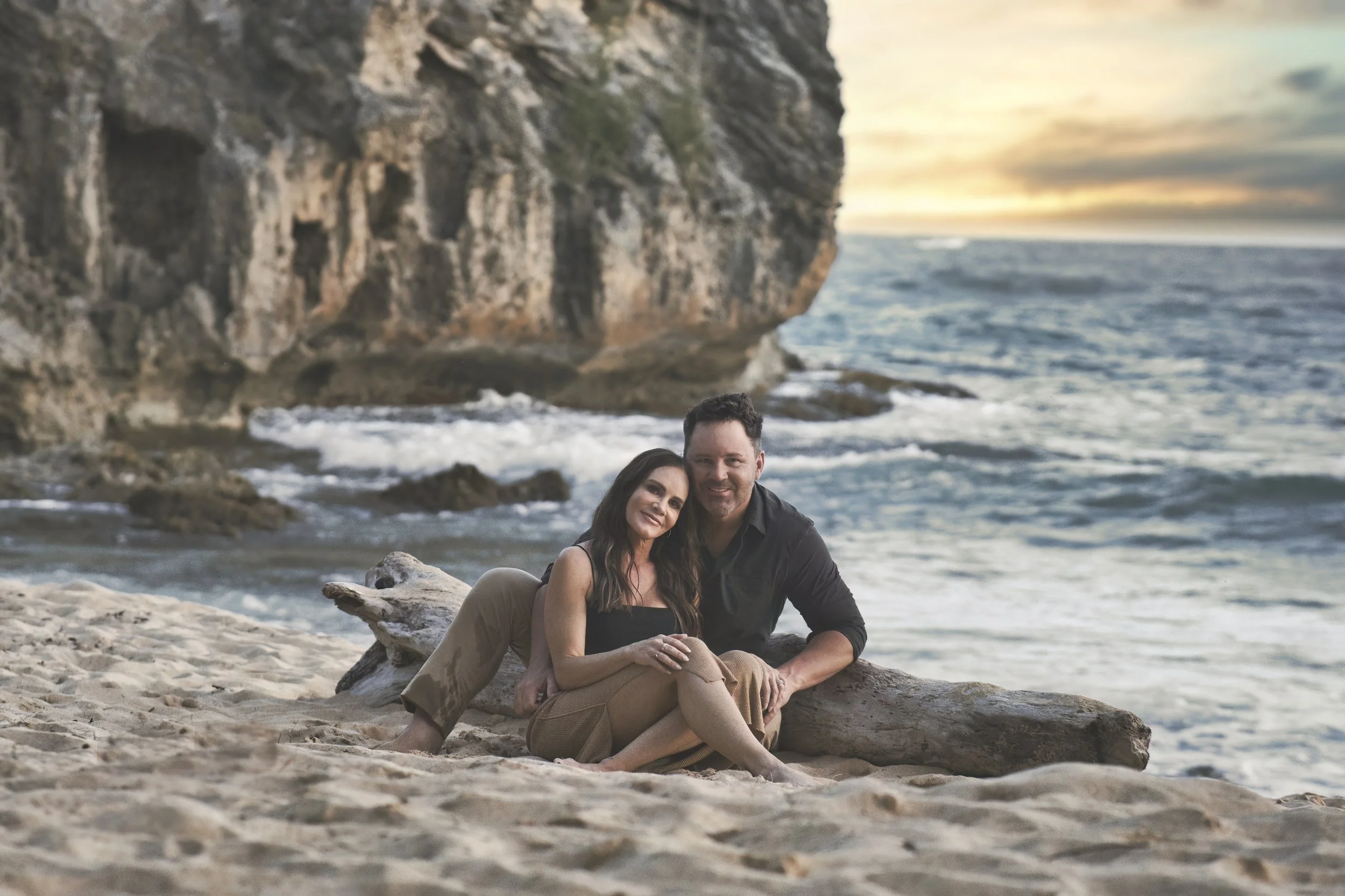 Relaxed couple sitting on driftwood with the ocean behind them at Shipwreck Beach, Kauai.