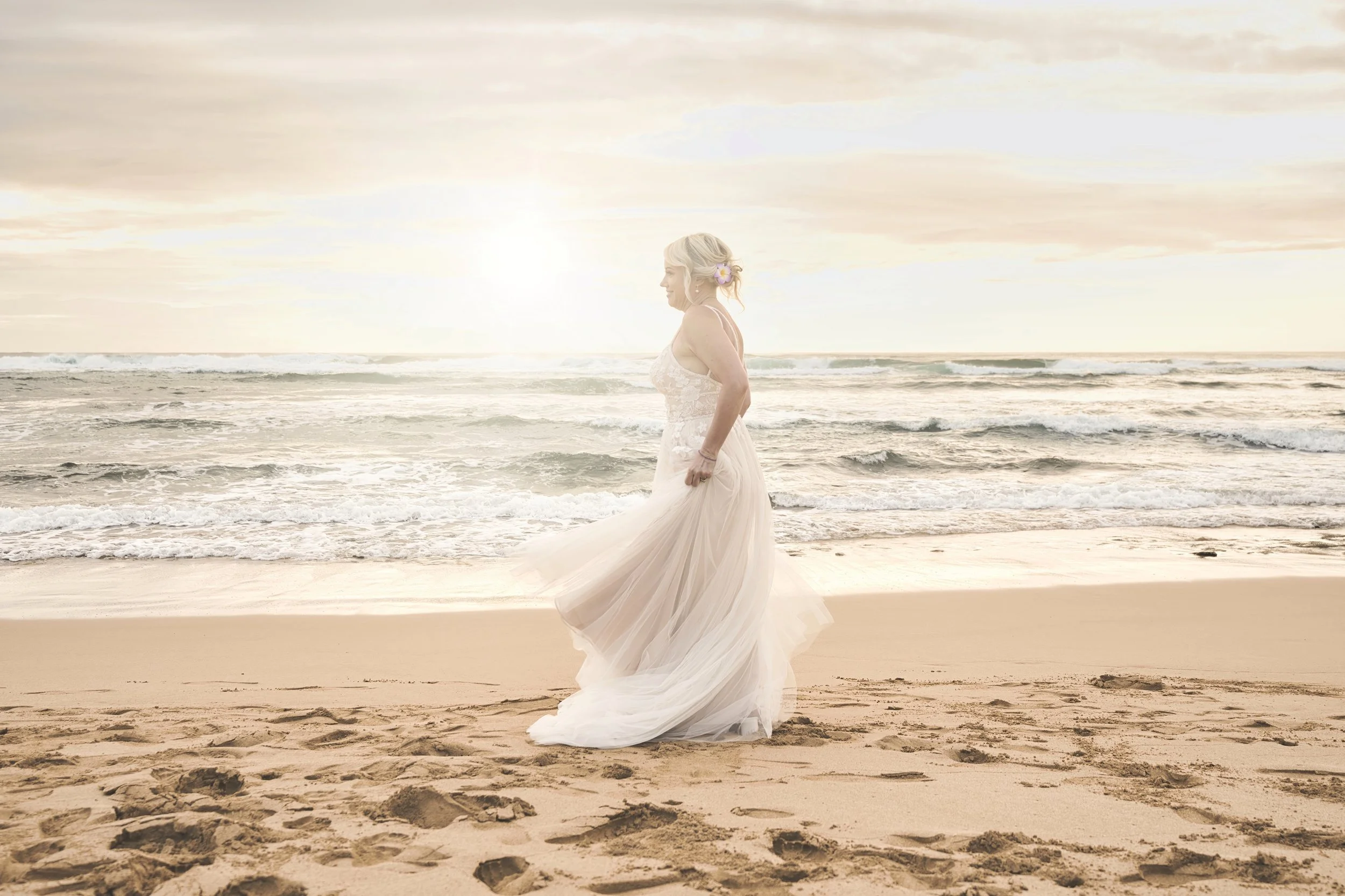 Bride in a flowing white gown standing alone on a Kauai beach in soft evening light.