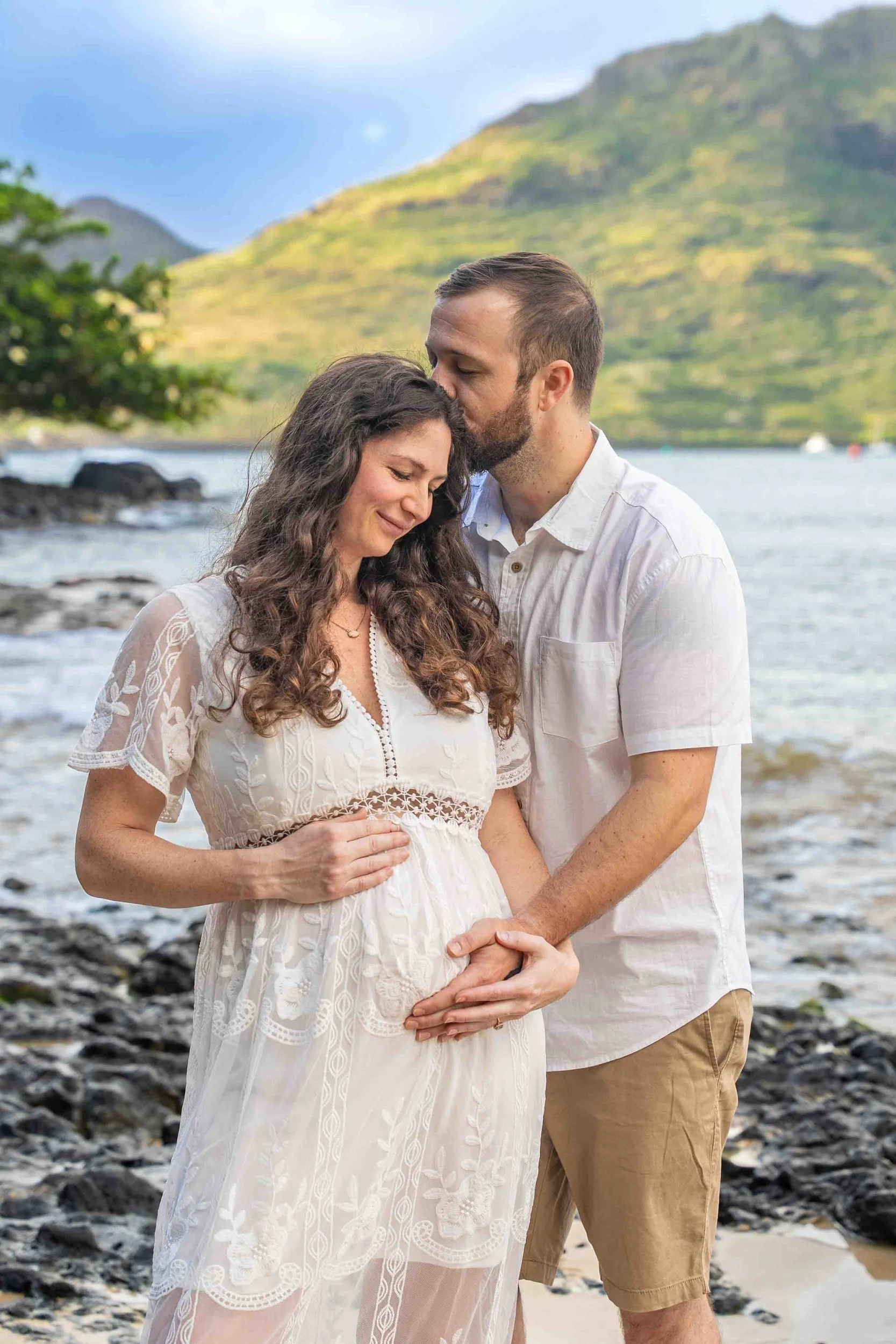 Expecting couple kissing while cradling the baby bump on a Kauai shore.