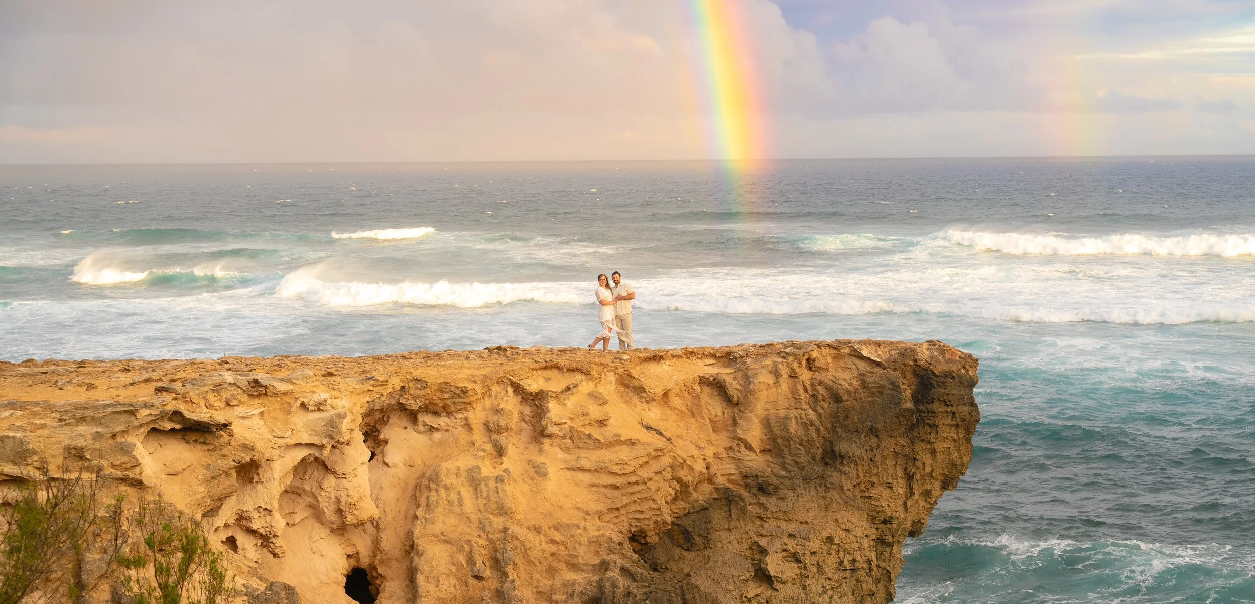 Couple embracing on a dramatic ocean cliff in Kauai beneath a rainbow.