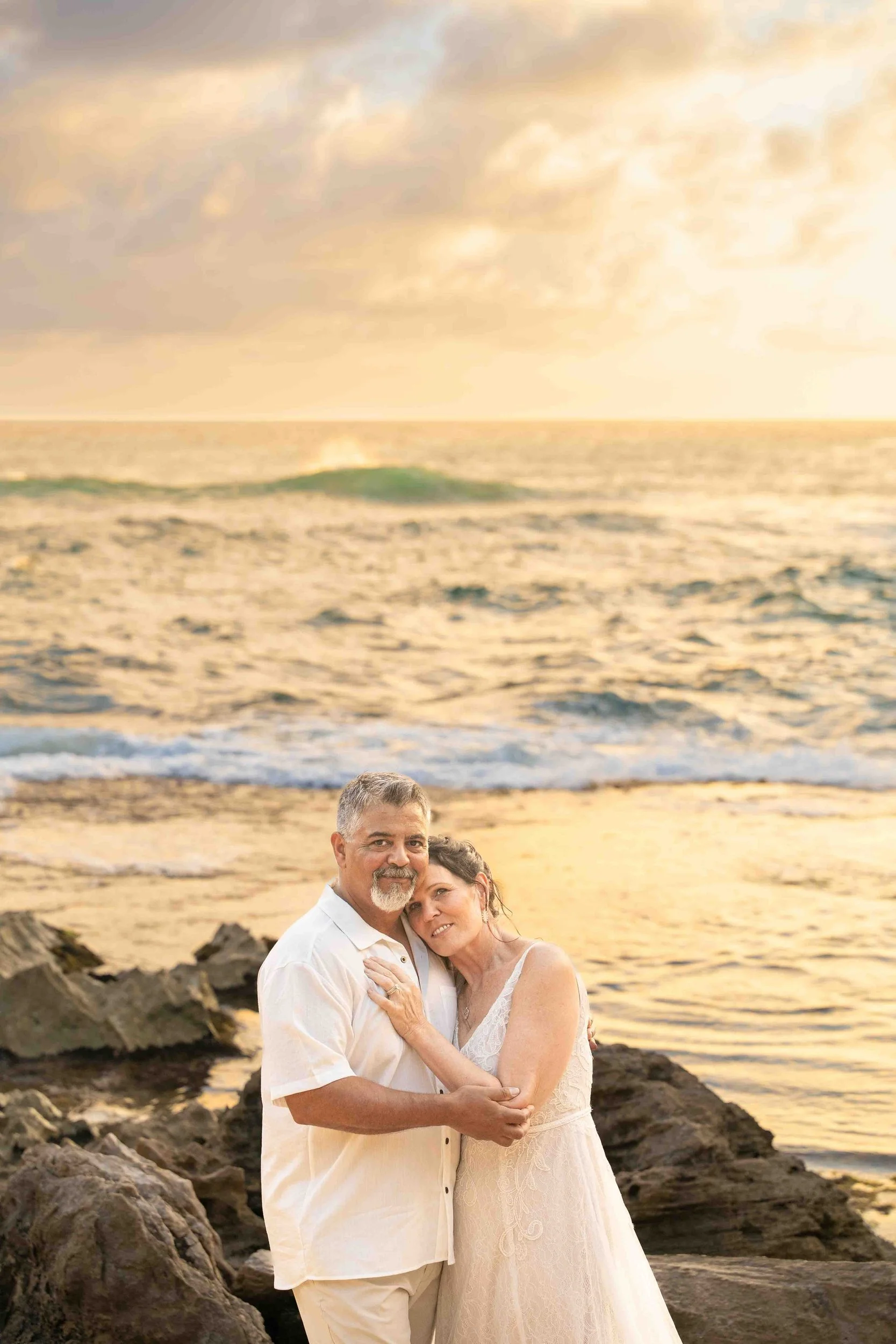 Older couple embracing on the rocky shore at sunset at Shipwreck Beach, Kauai.