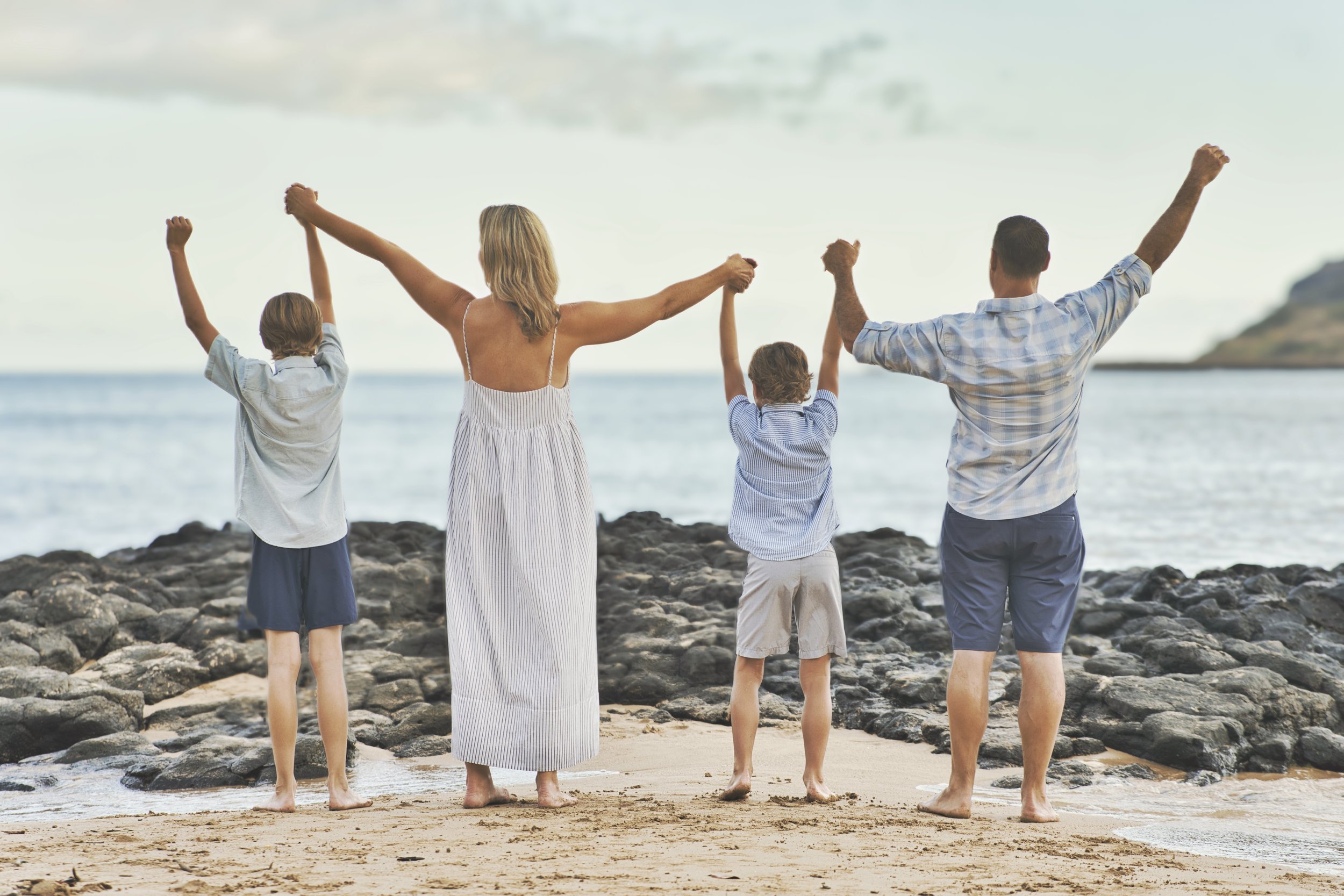 Family of four raising their hands toward the ocean on a rocky Kauai beach.